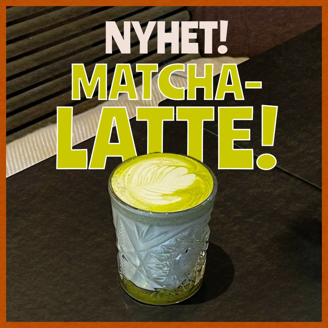 En matcha-latte i ett glas med ett vitt mönster, på en mörk bordsyta.