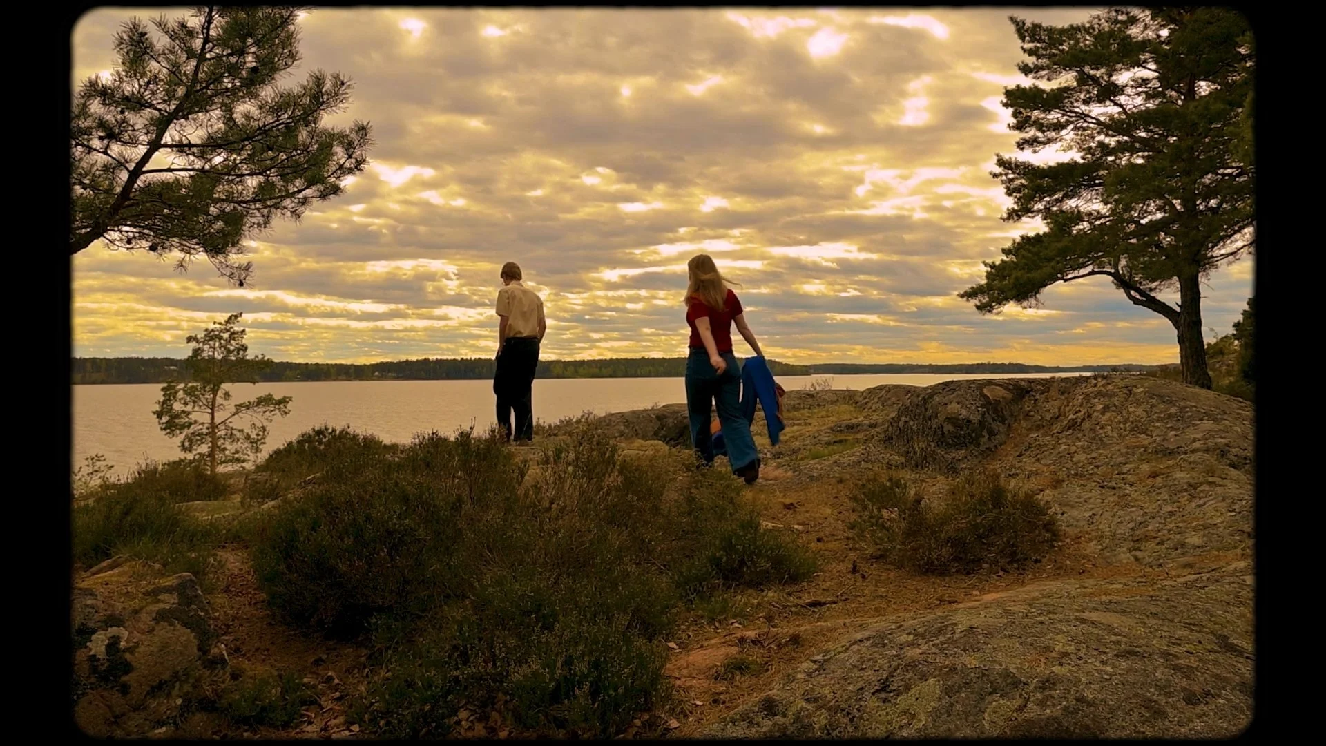 Två personer står på en klippa vid en sjö under en molnig himmel, omgivna av träd och buskar, vid skymning.
