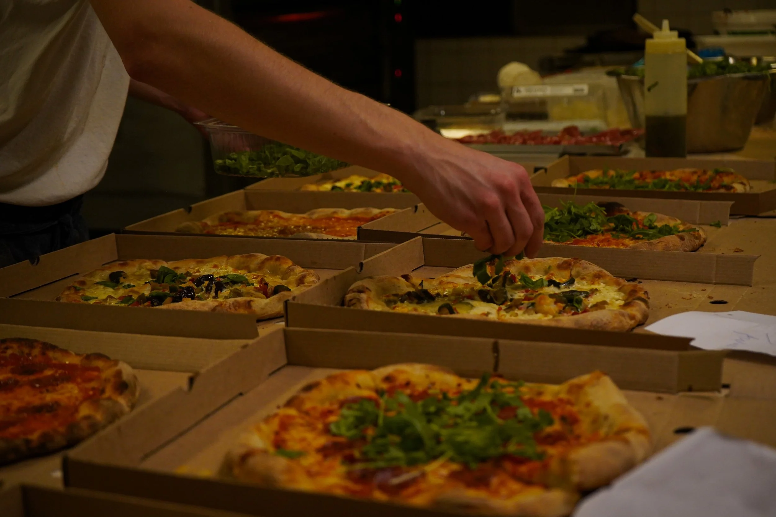 En person plockar upp pizza från en pappförpackning på en pizza-beredningsbänk, omgiven av flera pizzor med olika toppings.