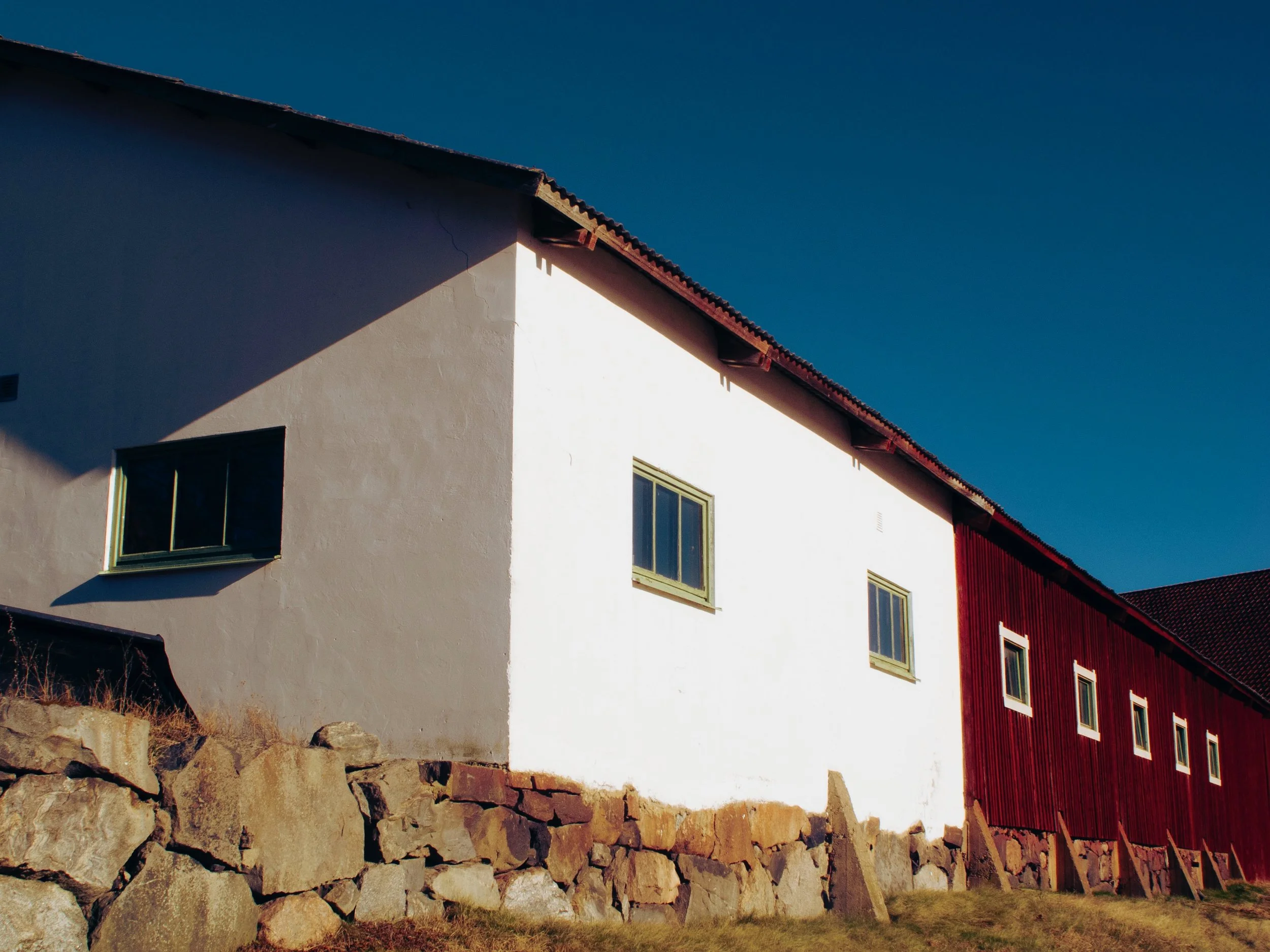 En vit och en röd byggnad med flera fönster under en klar blå himmel, med en stenmur i förgrunden.