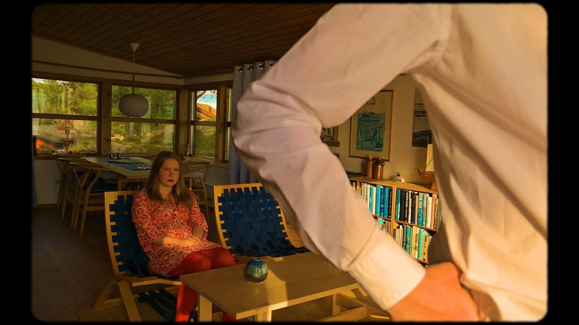 En ung kvinna sitter på en stol inomhus med armarna korsade. En person i vit skjorta står framför henne med ryggen mot kameran. Rummet har stora fönster med utsikt över gröna träd och ett hus utanför. På väggen finns konstverk, och ett bokhylla med mycket böcker.