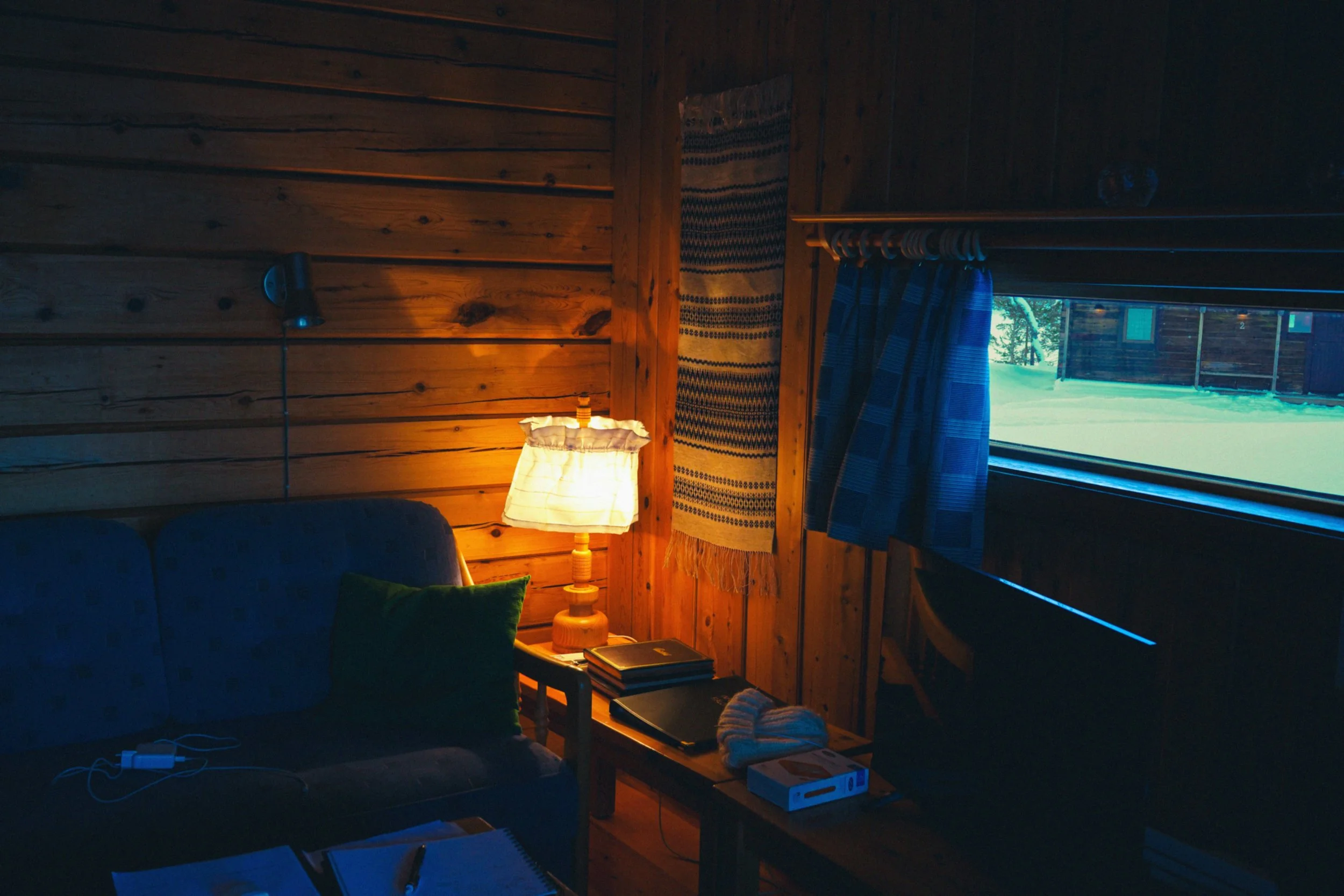 Sakrist vägg av träpanel, med en vägglampa, en pågående lampa, en bokhylla med böcker och kläder, ett fönster med blå randiga gardiner och utsikt över en snöig utomhusmiljö.