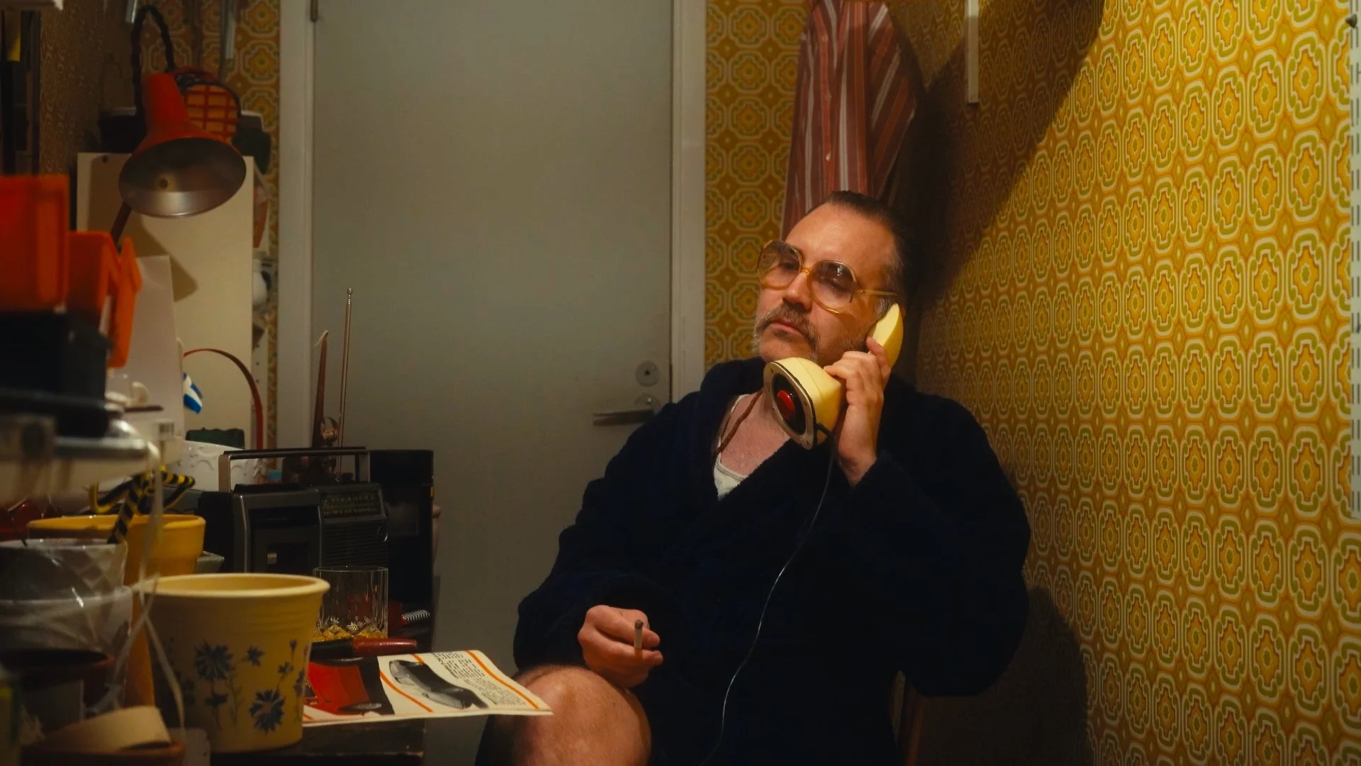 Man med skägg och solglasögon som sitter i ett hörn, pratar i en gul telefon. Runtomkring finns kaffekoppar och olika småsaker på ett bord, och väggarna är täckta av mörk orange och gul mönstrad tapet.