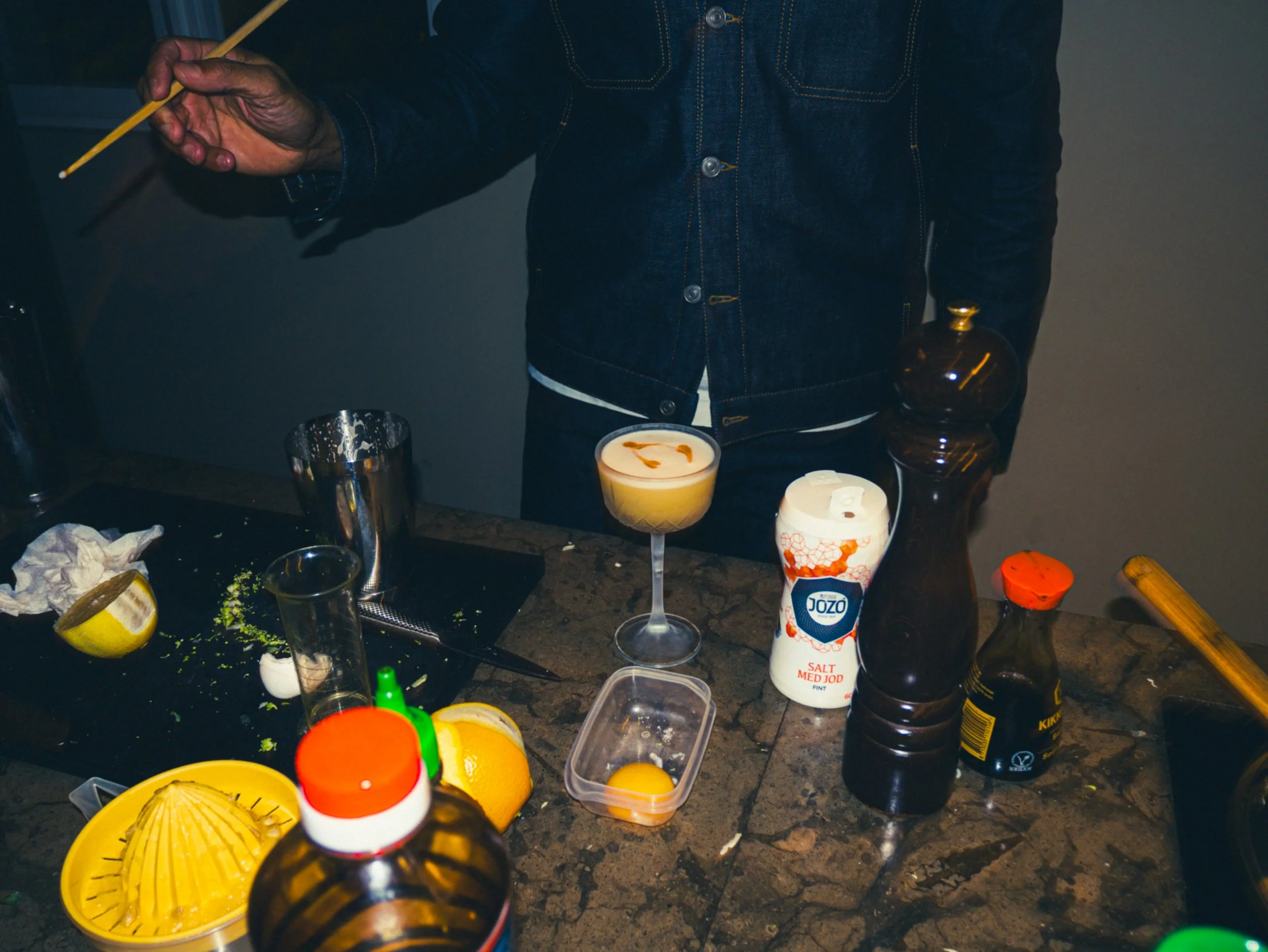 En person i mörk denimjacka står vid ett bord med olika köks- och bakverk, inklusive en cocktailglas med en drink, citrusfrukter, ägg, en kryddflaska, en skål och köksredskap.