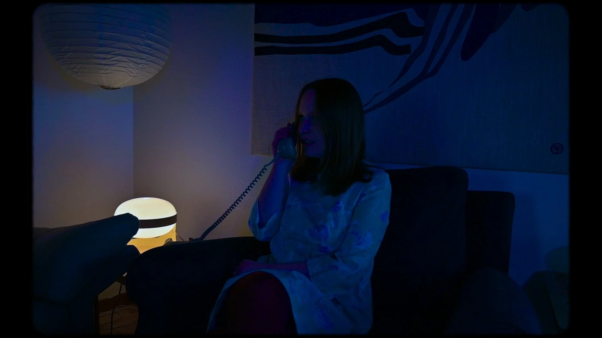 En ung kvinna sitter i ett vardagsrum på kvällen, pratar i telefonen. Rummet är mörkt, upplyst av ett nattduksbordsljus och en vägglampa. Det finns en väggdekoration i bakgrunden.