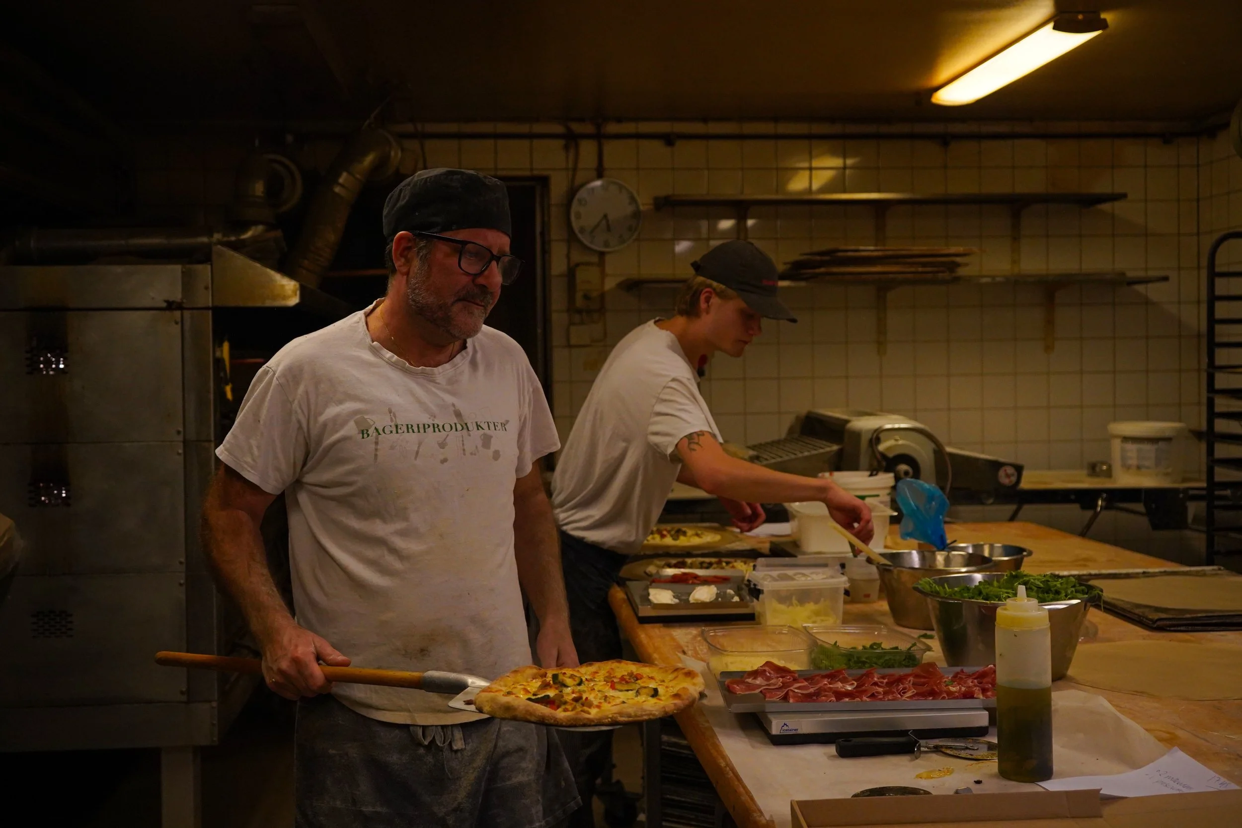 Två män i ett kök, den äldre känner eller gör något medan den yngre skär eller förbereder mat, omgivet av ingredienser och köksutrustning.