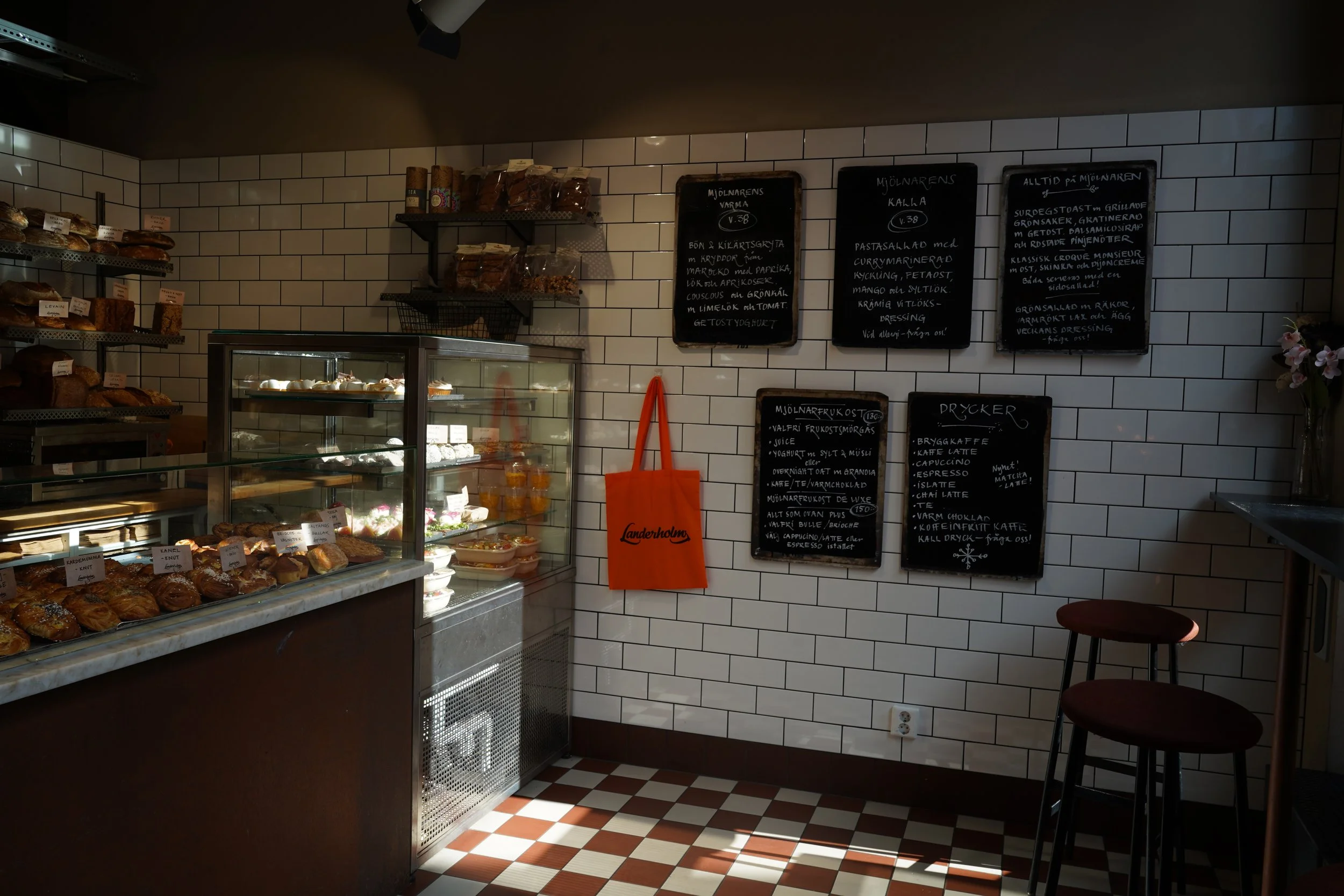 Interiör av ett kafé eller bageri med väggar klädda i vita glaserade tegelstenar. En glasmonter fyllt med bakverk, framför en vägg med svartkritstaväggar där det finns handskrivna menyer. Till vänster syns en hylla med olika bröd. Det finns ett rött tygväskorhängband med texten 'Landerholm' i orange.