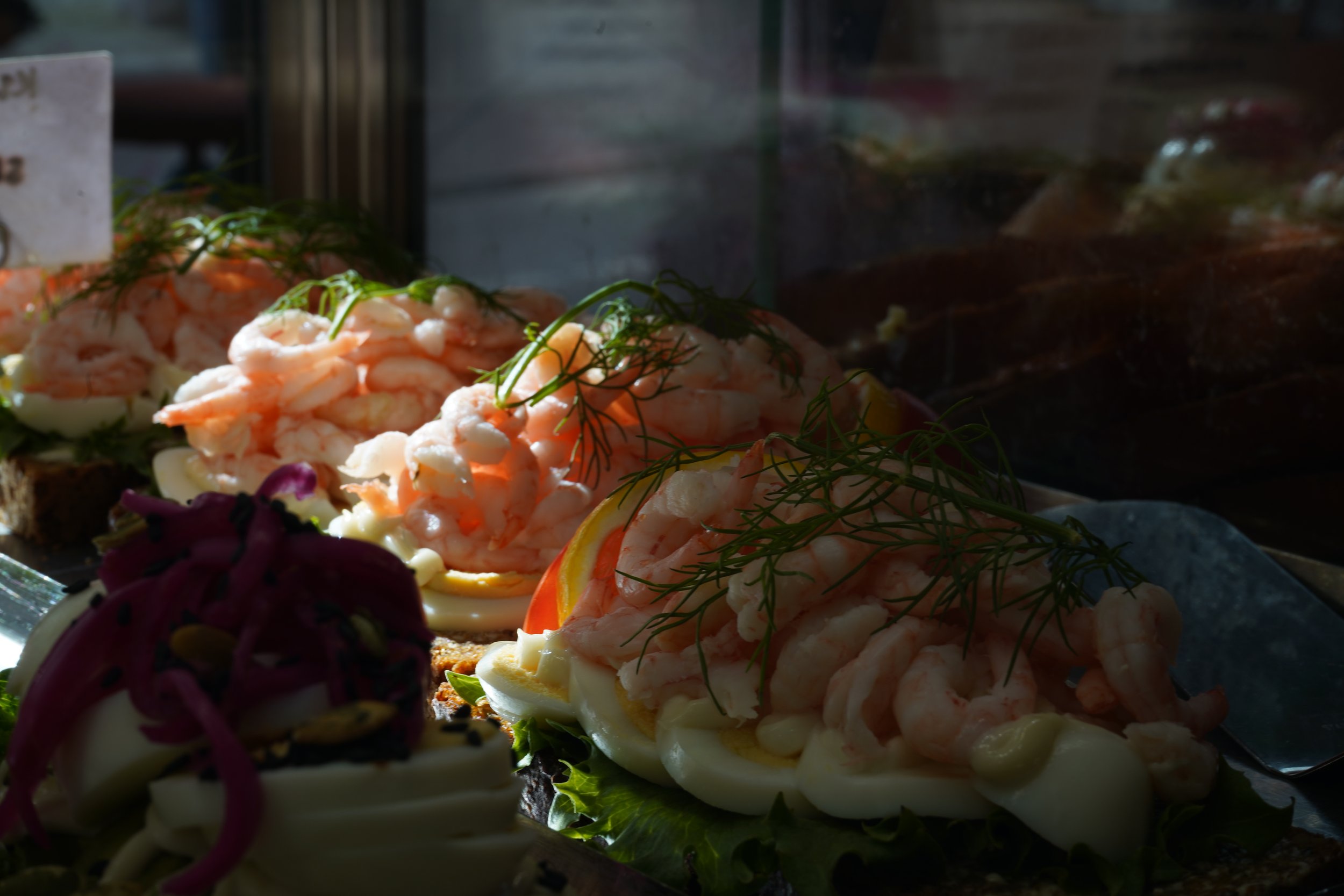 Flera smörgåsar med räkor, ägg, dill och grönsaker på ett bord vid ett fönster.