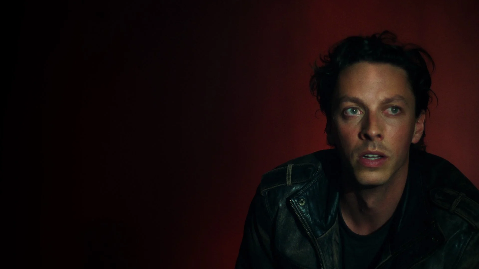 Close-up of a man with black curly hair and green eyes, wearing a black leather jacket, against a red background.