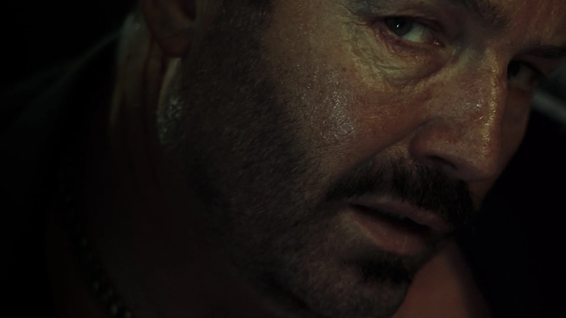 Close-up of a man's face with dark hair, beard, and mustache, with a focus on the right side of his face, showing his eye, nose, and part of his mouth, in a dimly lit environment.