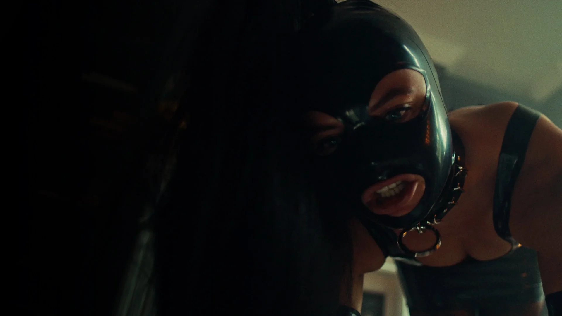 A person wearing a black helmet and face mask, with visible eyes and facial expression, appears to be leaning on or against a dark surface in a dimly lit environment.