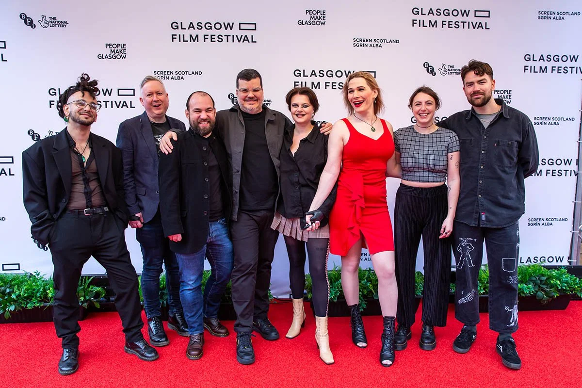 Group of eight people standing on a red carpet at the Glasgow Film Festival, posing for a photo with a white backdrop featuring event logos and text.