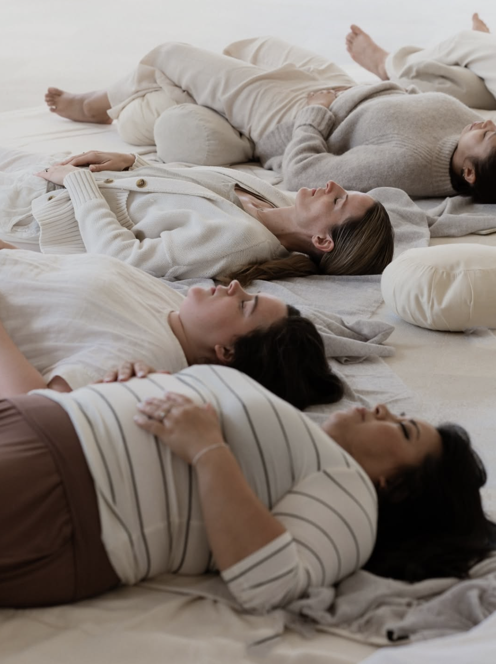 Five women lie on their backs on a bed with light-colored sheets, resting with eyes closed, wearing casual neutral-toned clothing, in a peaceful scene.