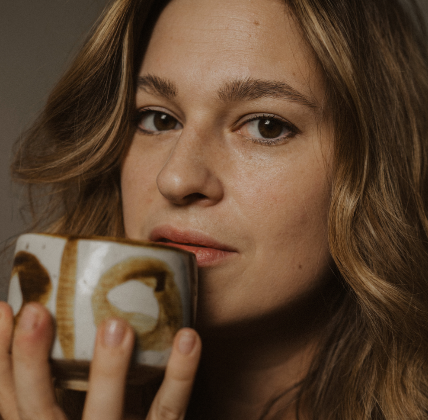 A young woman with wavy brown hair holding a coffee mug near her face, looking directly at the camera.