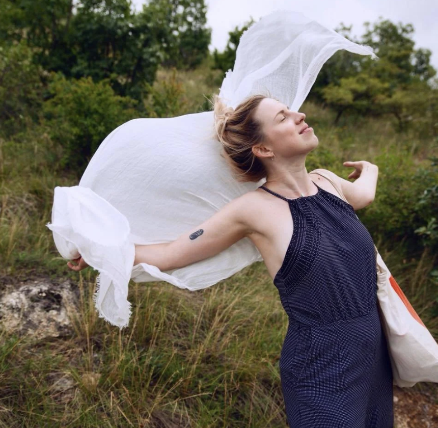 A woman with short hair, wearing a sleeveless dark blue dress, stands outdoors in a grassy area with trees, holding a large white cloth that is flowing in the wind behind her.