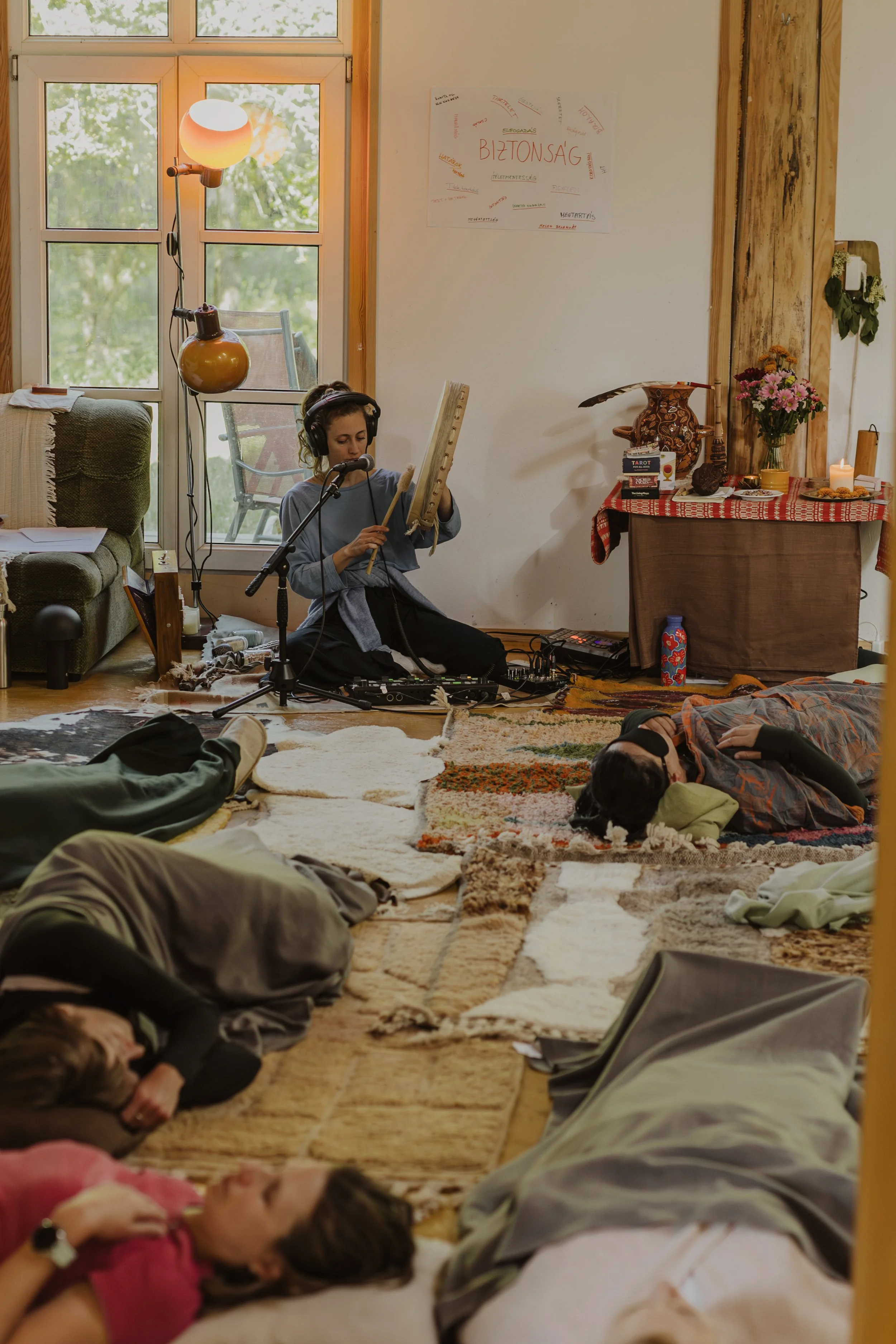 Group of children sleeping on a group of blankets and rugs on the floor, with some people lying on the ground and others sitting or kneeling. A woman in the background is sitting on the floor with headphones and a percussion instrument, near a window and a small altar with flowers and candles.