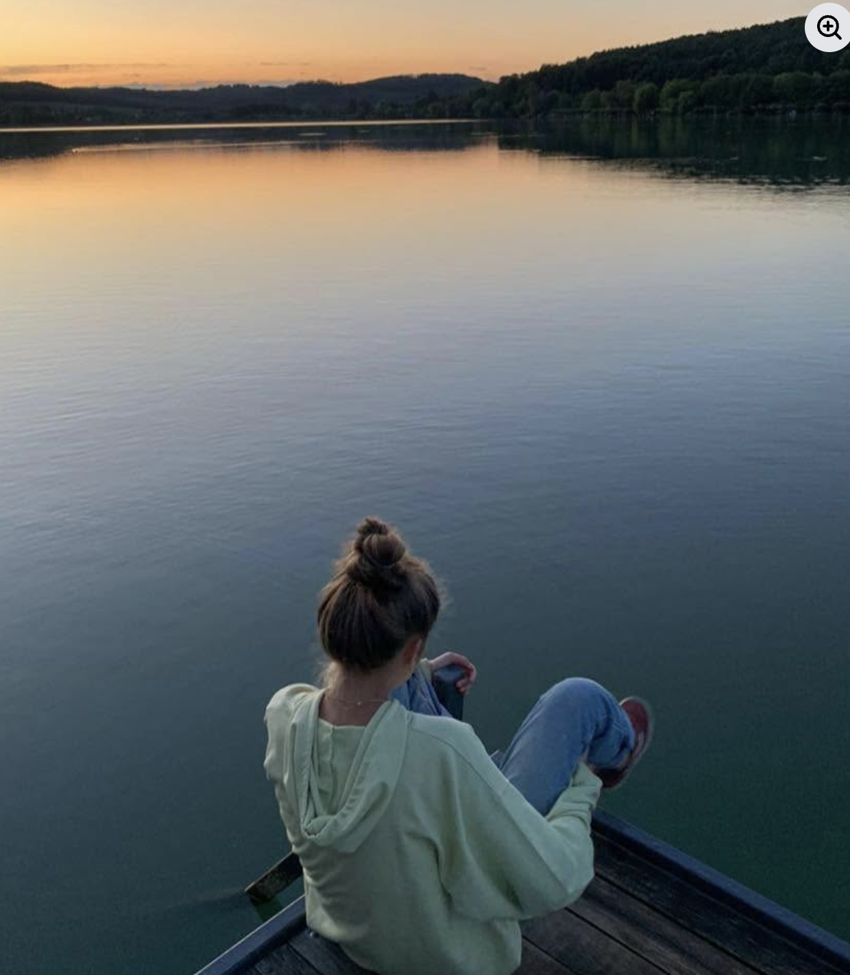 A person sitting on a dock by a calm lake during sunset, with hills and trees in the background.