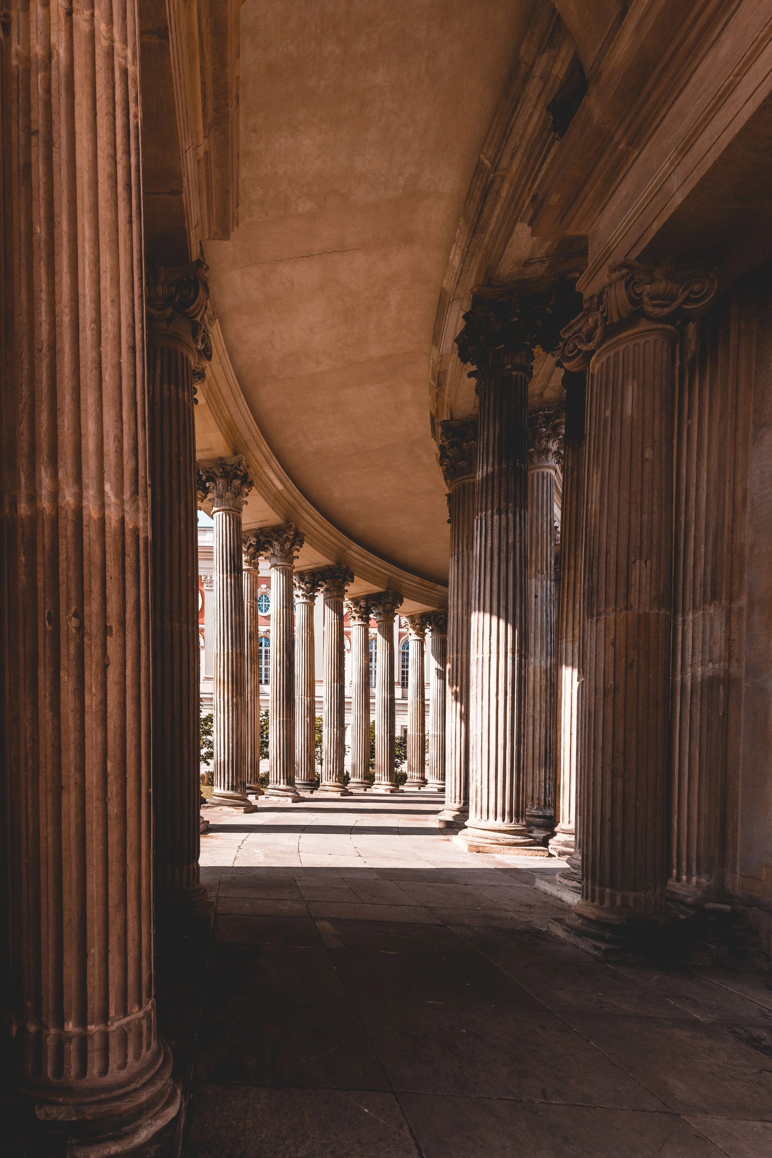 Columned corridor with classical architecture, showing tall, ornate columns and a curved ceiling, with sunlight casting shadows.