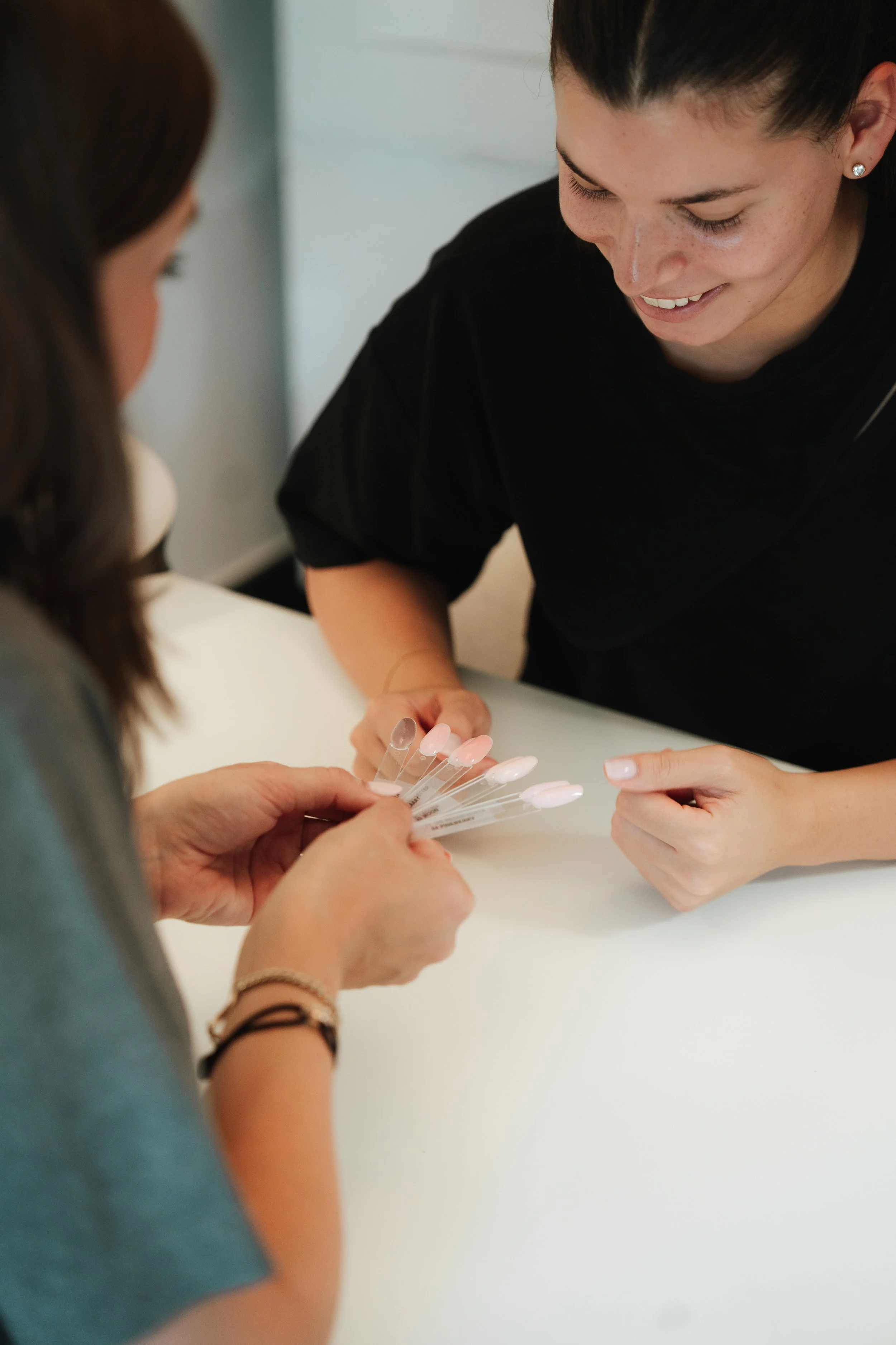 Close-up of two women exchanging nail polish samples at a salon table, one woman in a grey shirt and the other in a black shirt smiling.