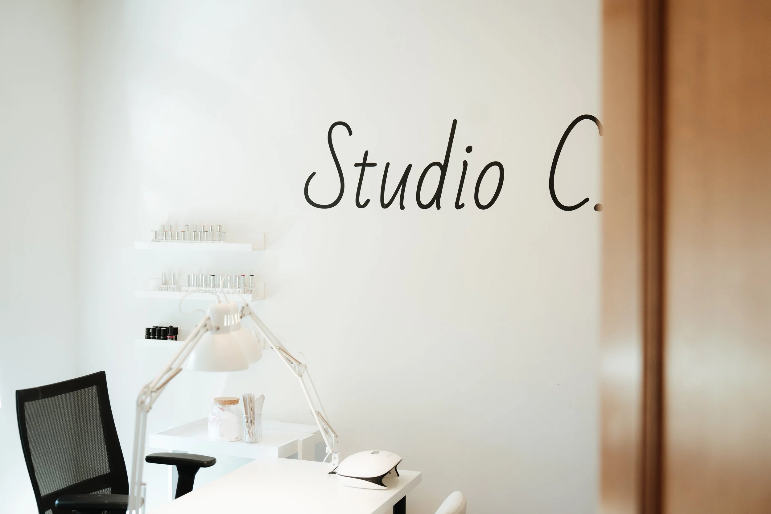 A reception or consultation area in a beauty or nail salon, with a white wall displaying the words 'studio C.' in black cursive. There are shelves with nail polish bottles, an adjustable desk lamp, a black chair, and a white table with items including a computer mouse and a jar with wooden sticks.