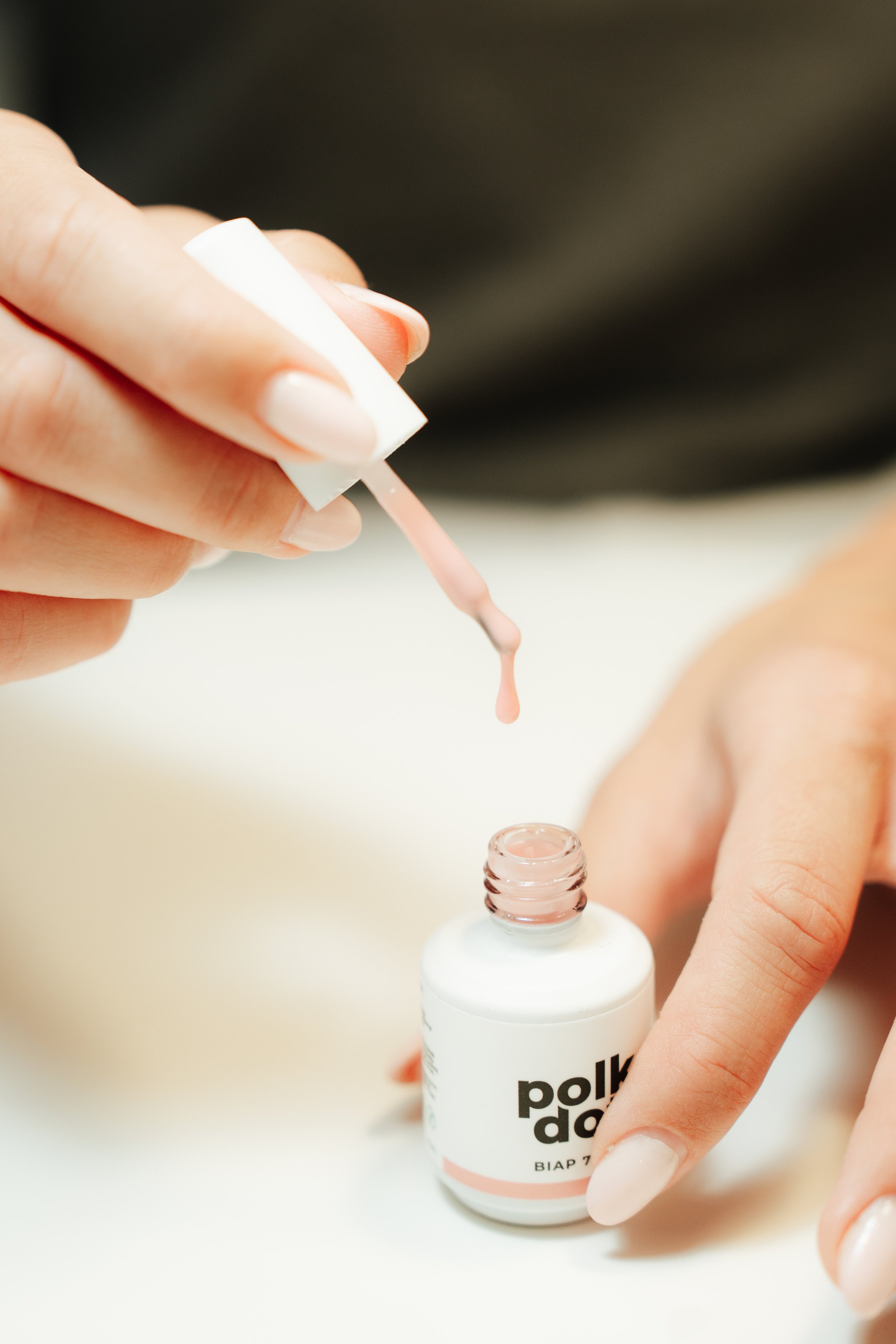 Close-up of a person's hand holding a bottle of polik dolk BISAP 7 liquid, with the other hand holding a cotton swab applying liquid to the person's fingernail. The background is blurred.
