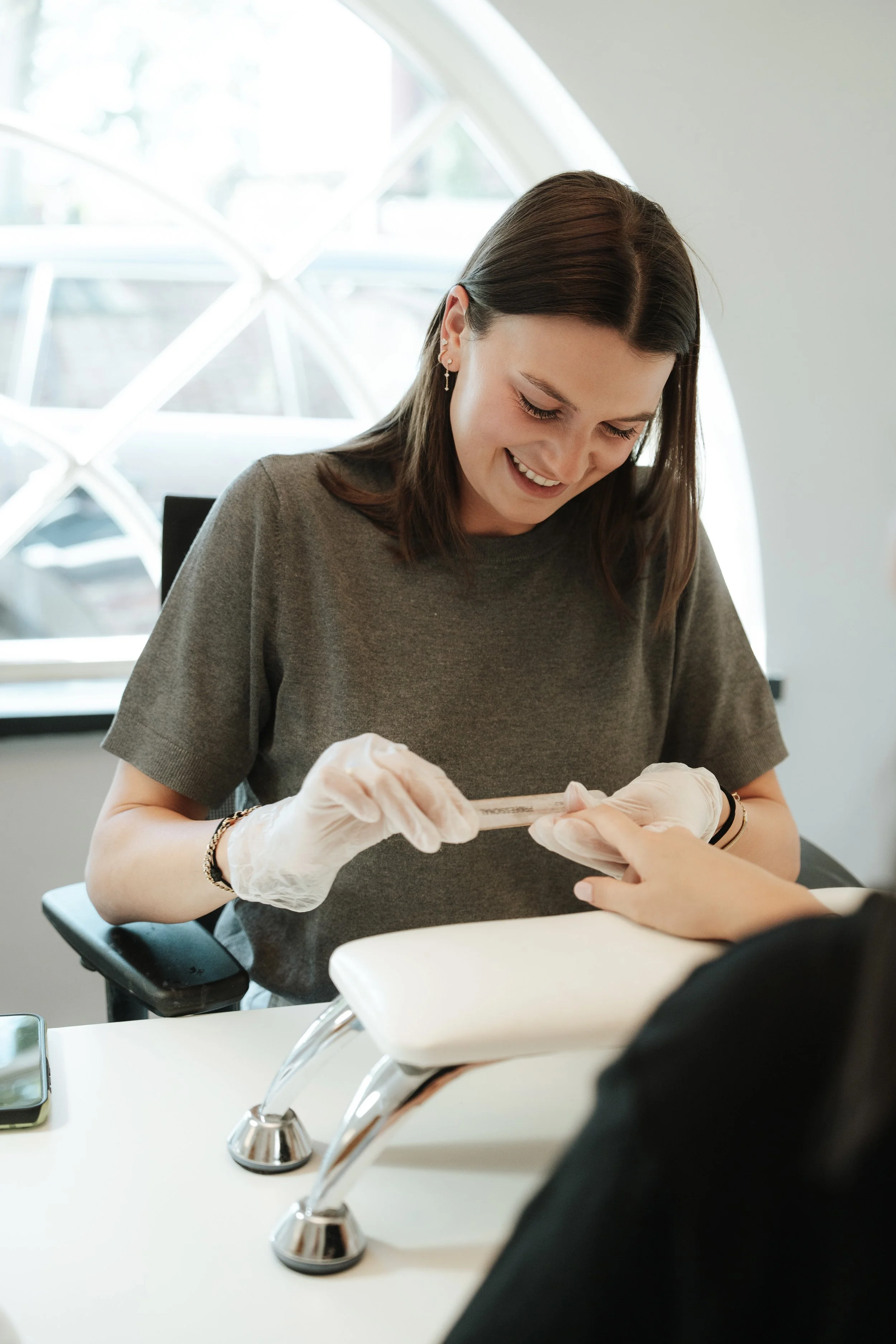 A woman smiling as she gets a manicure at a nail salon, wearing white gloves, with a manicurist's hand visible applying nail polish.