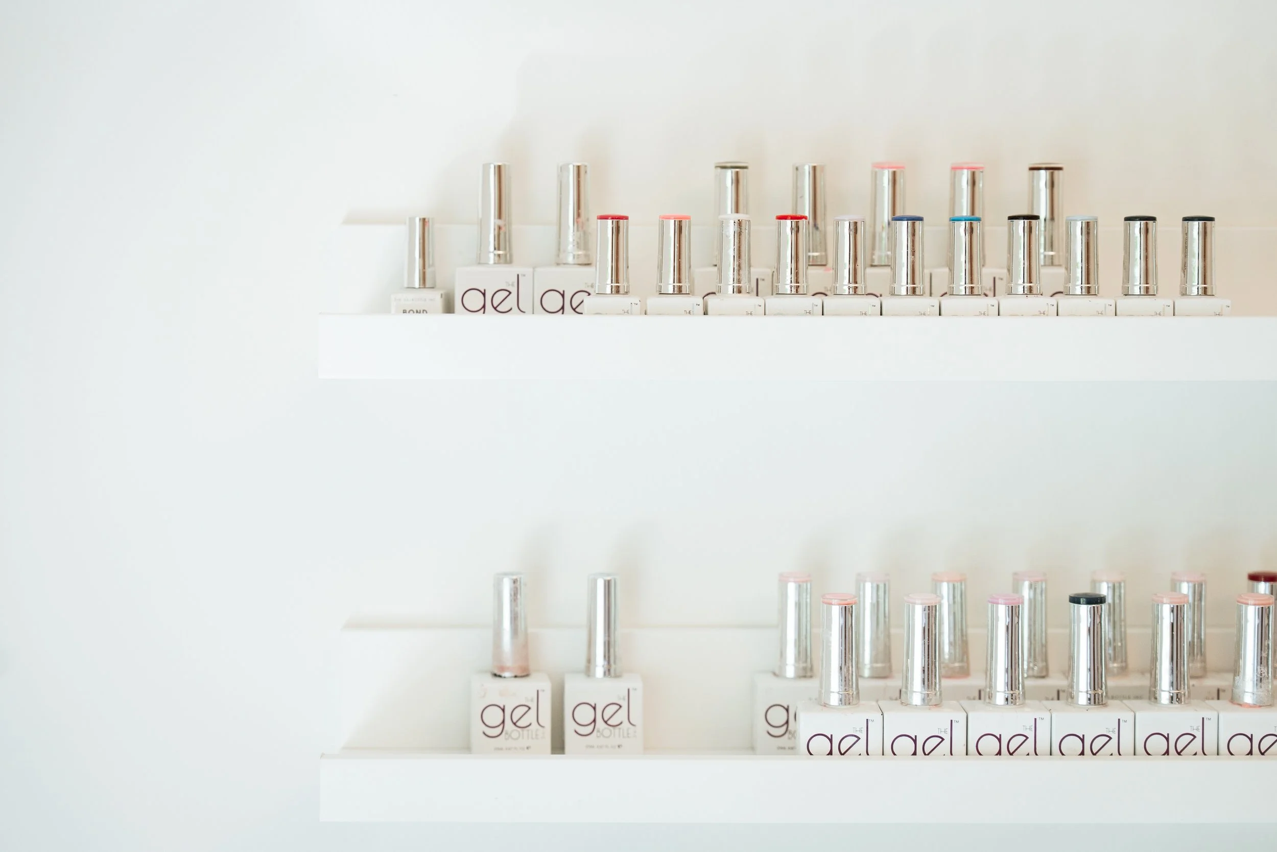 A display of nail polish bottles with metallic caps and labeled boxes of gel nail polish arranged in rows on white shelves.