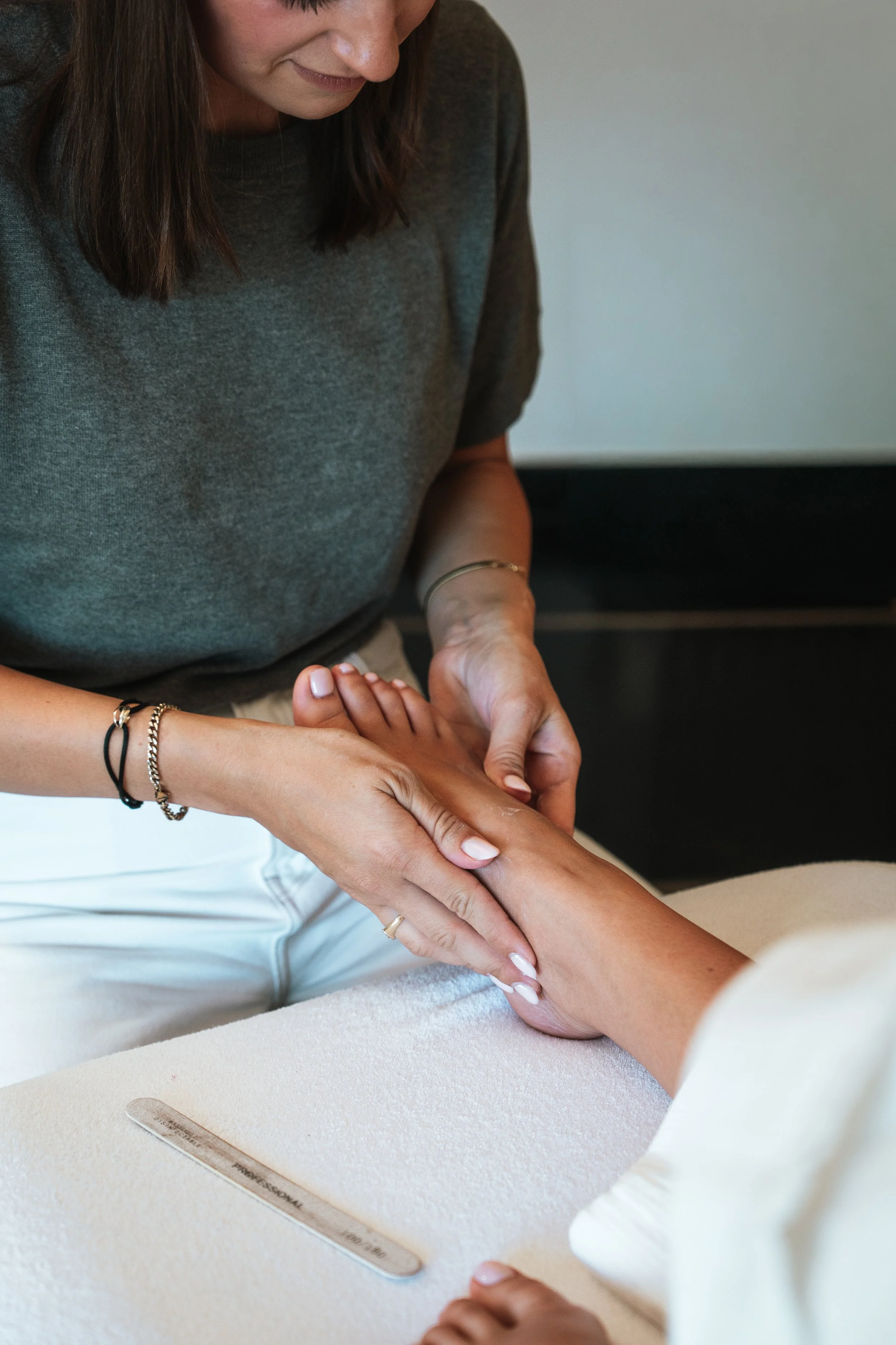A woman with long brown hair wearing a grey top is giving a manicure to another person's hand, which is resting on a soft white surface. A metal nail file with text is visible on the surface.
