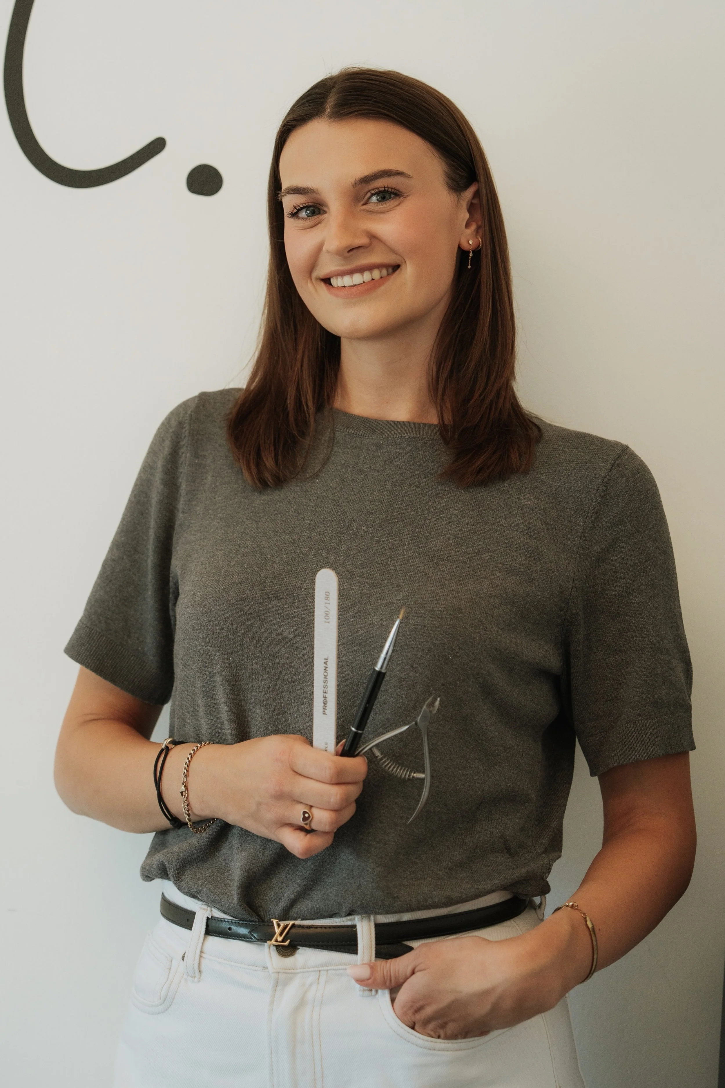 A young woman with long brown hair, smiling, wearing a gray t-shirt and white pants, holding tattoo needles and tools in her hand. She is standing against a white wall with a partial black graphic design.