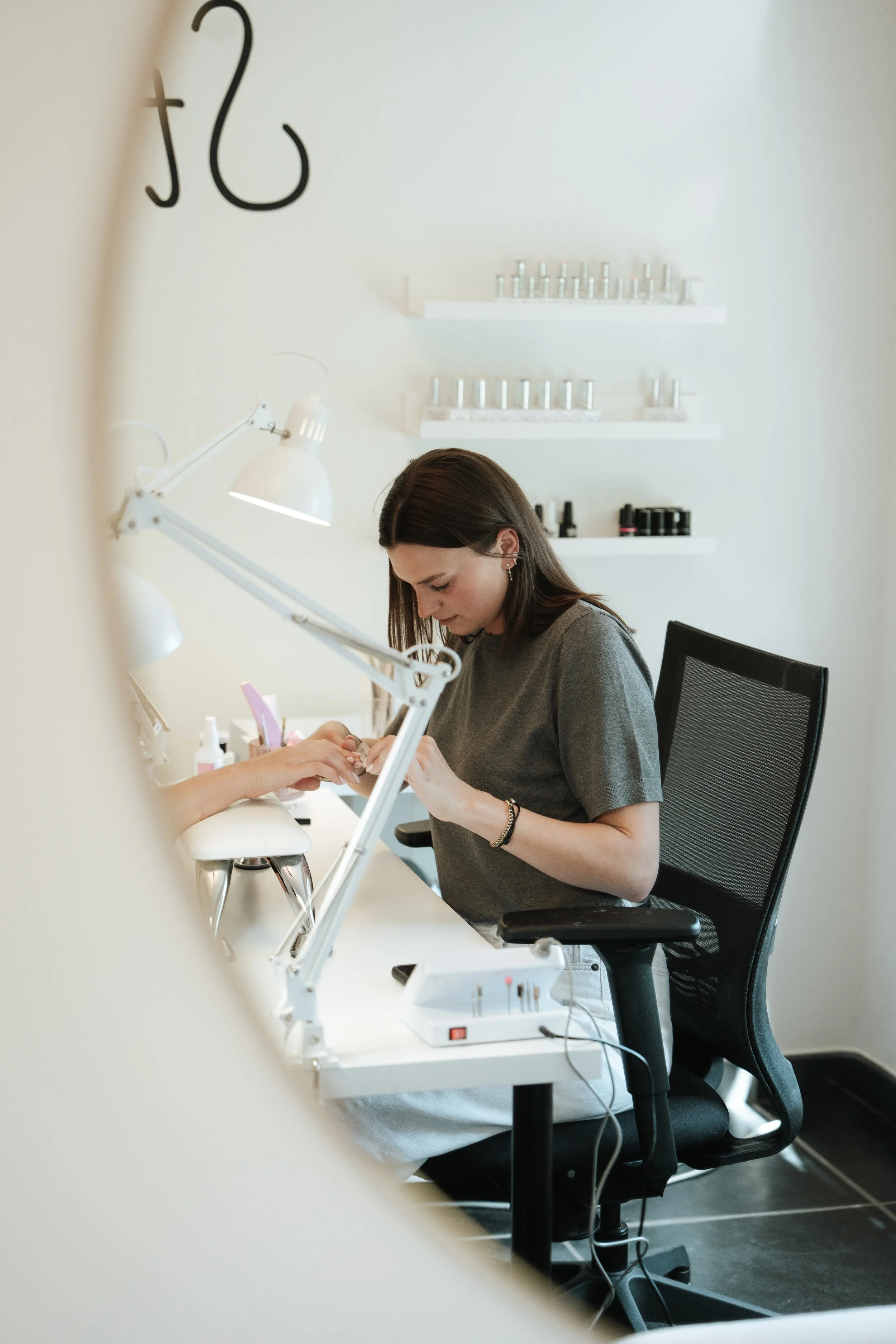 A woman with dark hair caring for a nail on a client's hand at a manicure station.