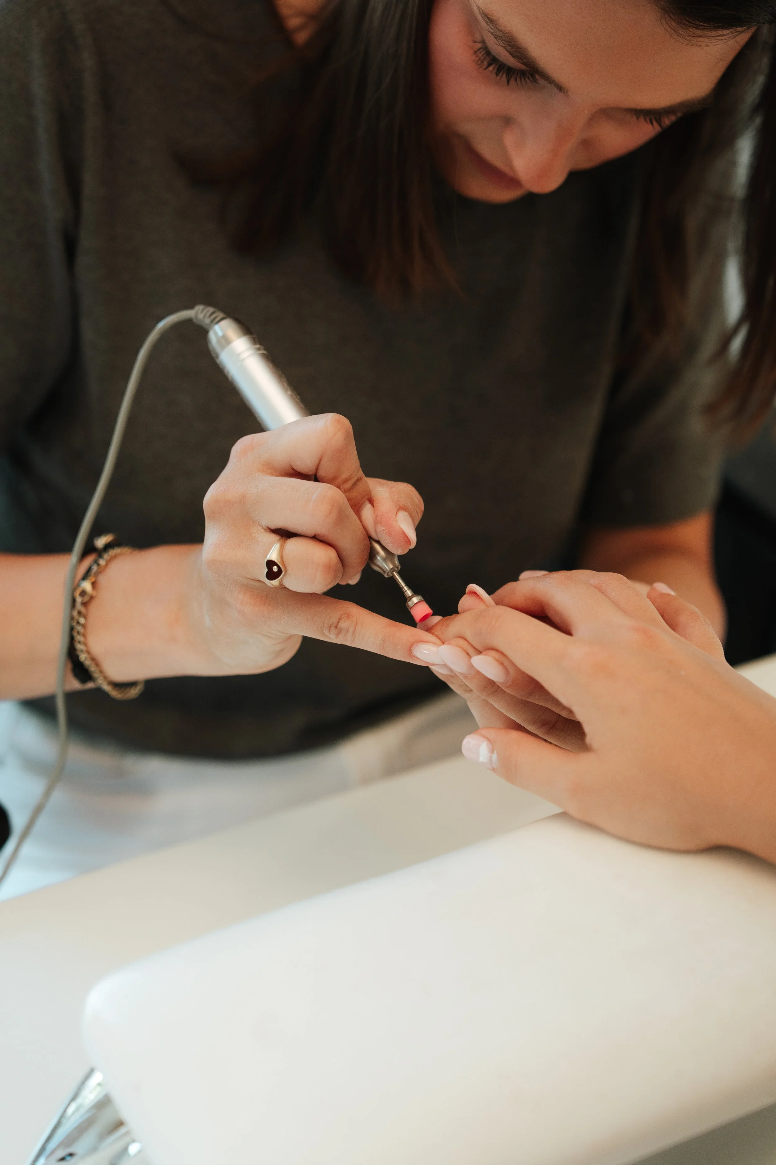 A woman with brown hair, wearing a dark gray shirt, is using a device to perform a manicure or nail treatment on another person's hand at a white table.