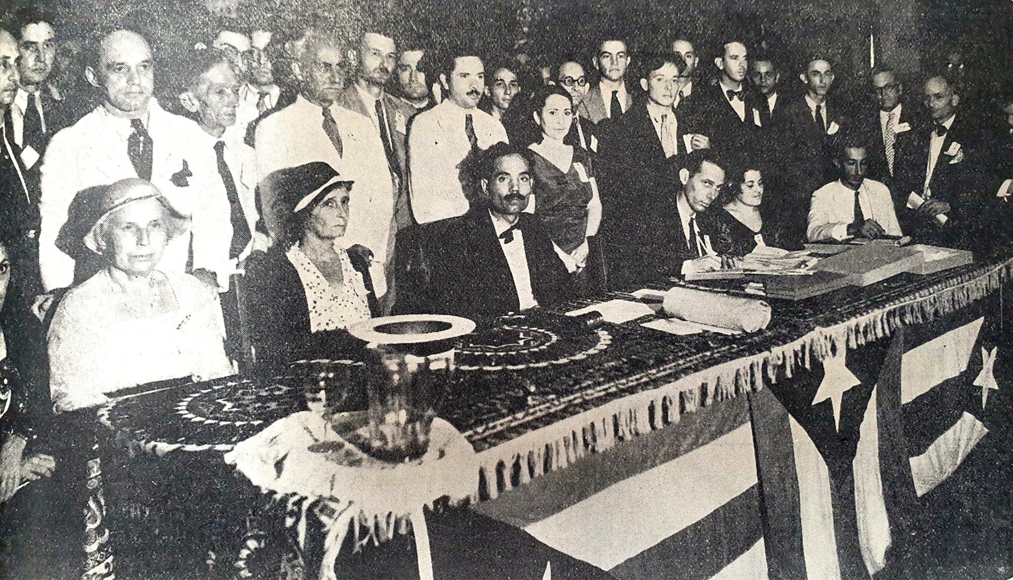 Black and white photograph of a formal gathering with people dressed in suits and dresses, some seated at a decorated table, and others standing behind. The scene suggests an official or celebratory event.