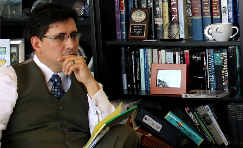 Man in glasses reading a book in a library with bookshelves behind him.
