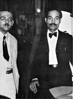 Two men dressed in formal attire, standing indoors. One man wears a tuxedo with a bow tie, and the other man wears glasses, a white shirt, and a dark tie.