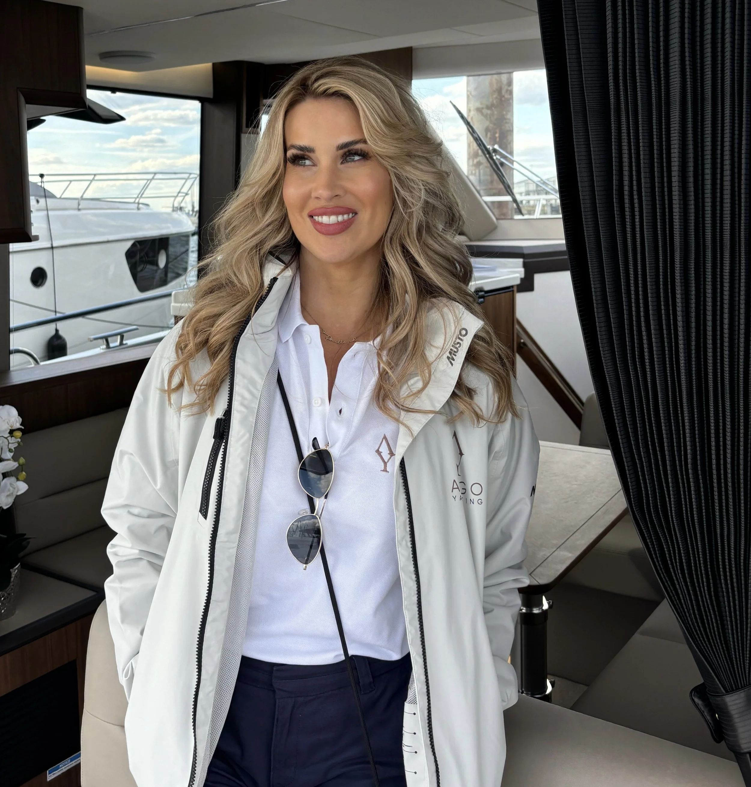 A woman with long wavy blonde hair, wearing a white jacket and dark pants, smiles and looks to her right inside a boat or yacht, with large windows and a view of another boat docked outside.