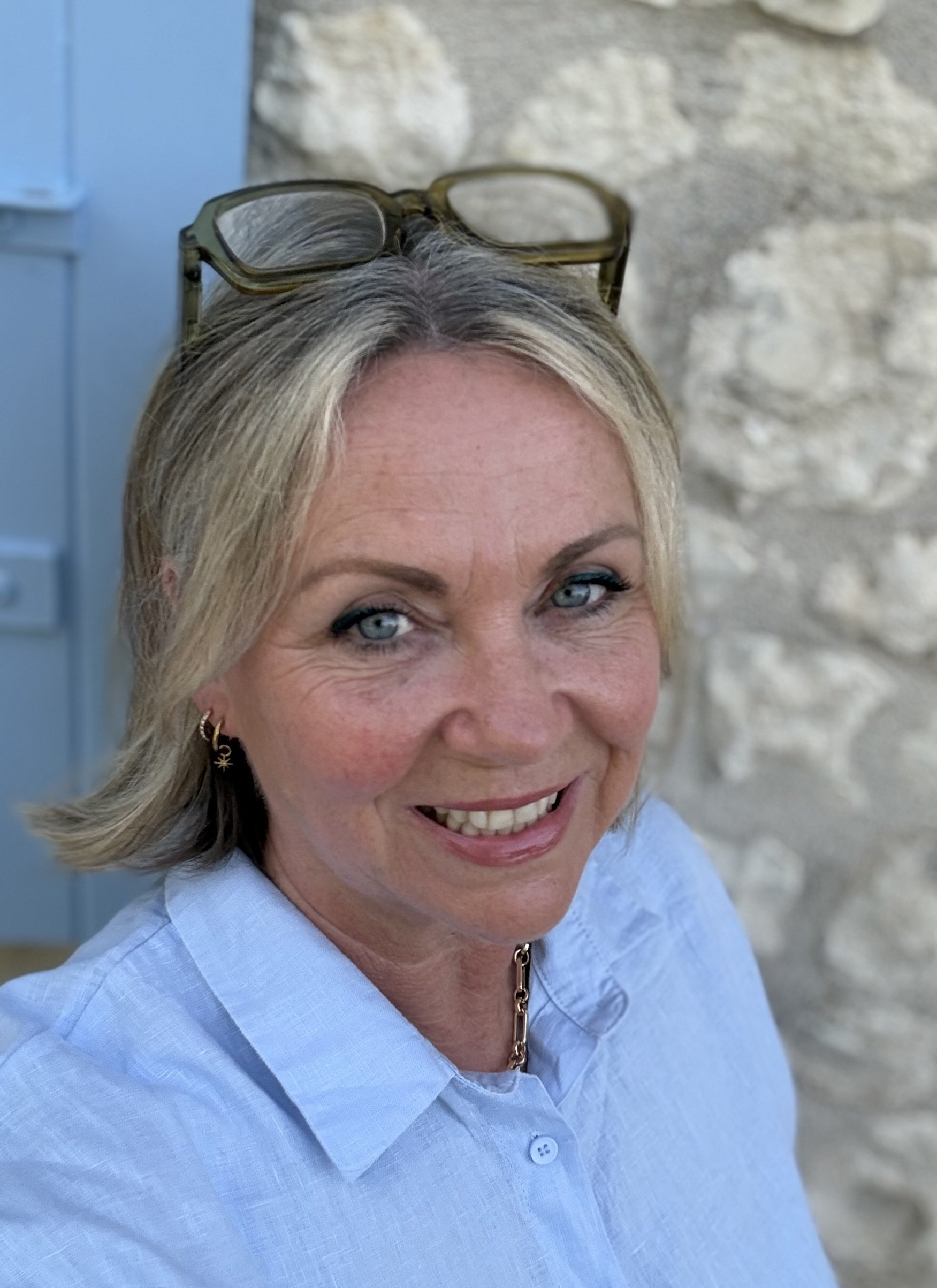Close-up of a smiling woman with blonde hair wearing glasses resting on her head, a light blue top, and a gold necklace, outdoors near a stone wall.