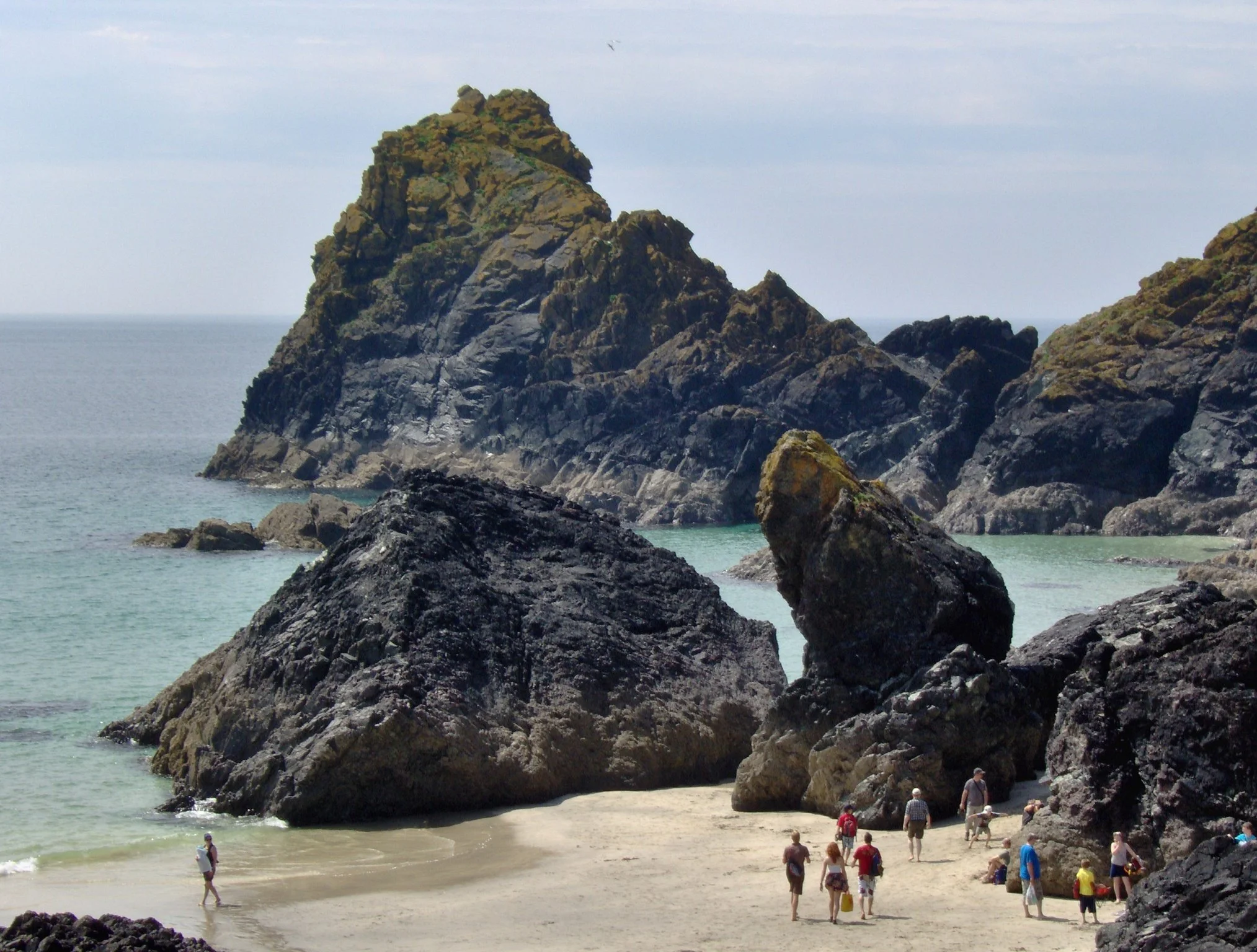 People walking along a sandy beach with large rocky formations in the background and the ocean in the distance.
