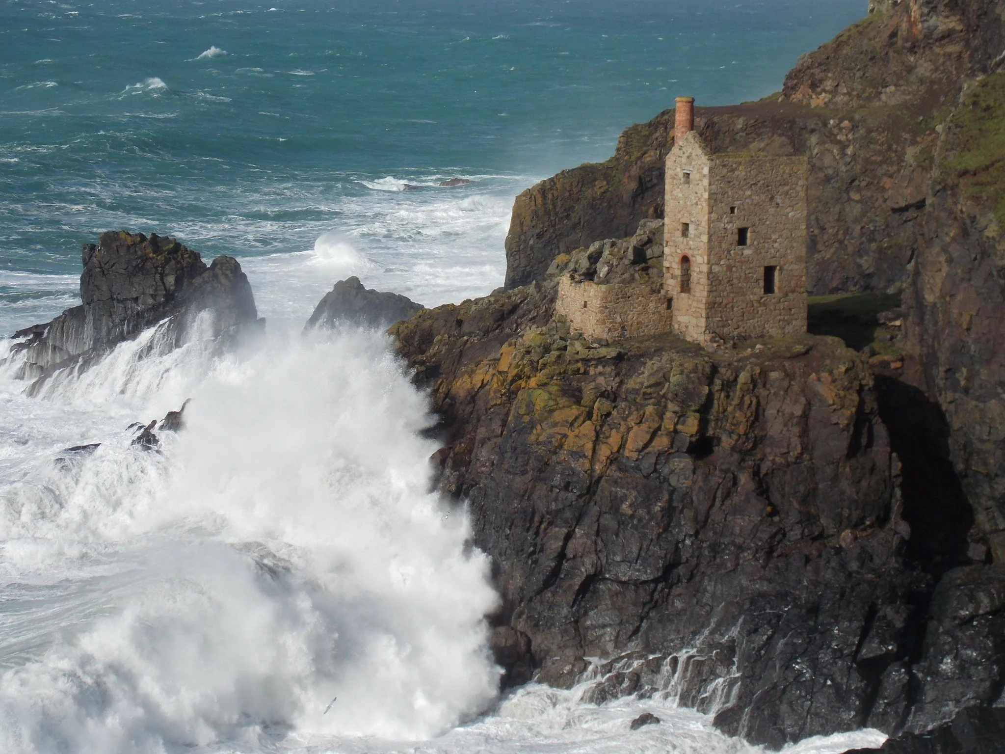 Cliffs along the coastline at Botallack with ruined engine houses and turbulent ocean waves crashing against rocks.