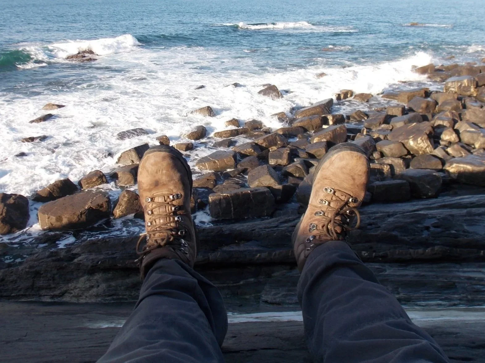 View of a person’s legs and feet in hiking boots resting on a rock ledge above a rocky shoreline with waves crashing against the rocks.