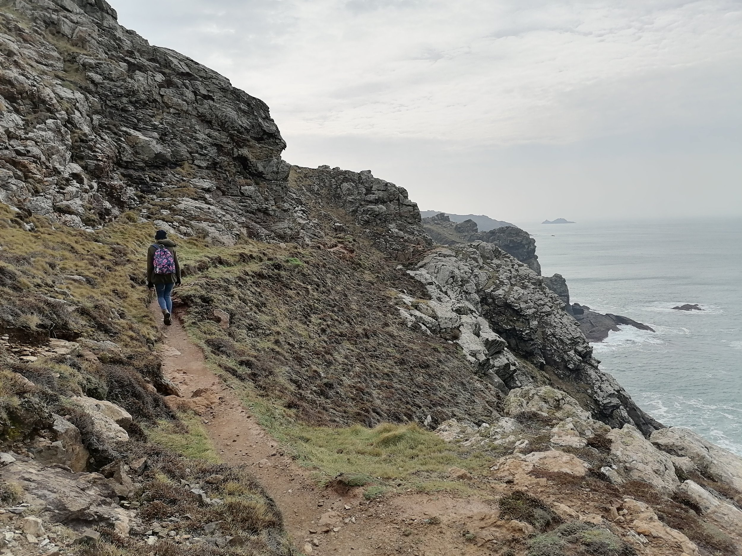 A person hiking along a narrow old miners path near Botallack on a coastal hillside with rocky terrain and sparse vegetation, overlooking the ocean with distant islands and an overcast sky.