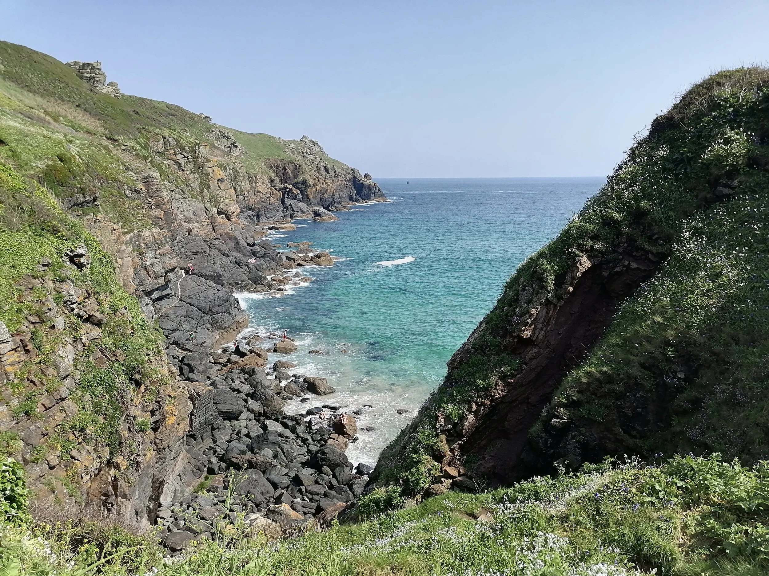 View of a rocky cove with lush green hills, blue ocean, and a sailboat in the distance on a clear day.