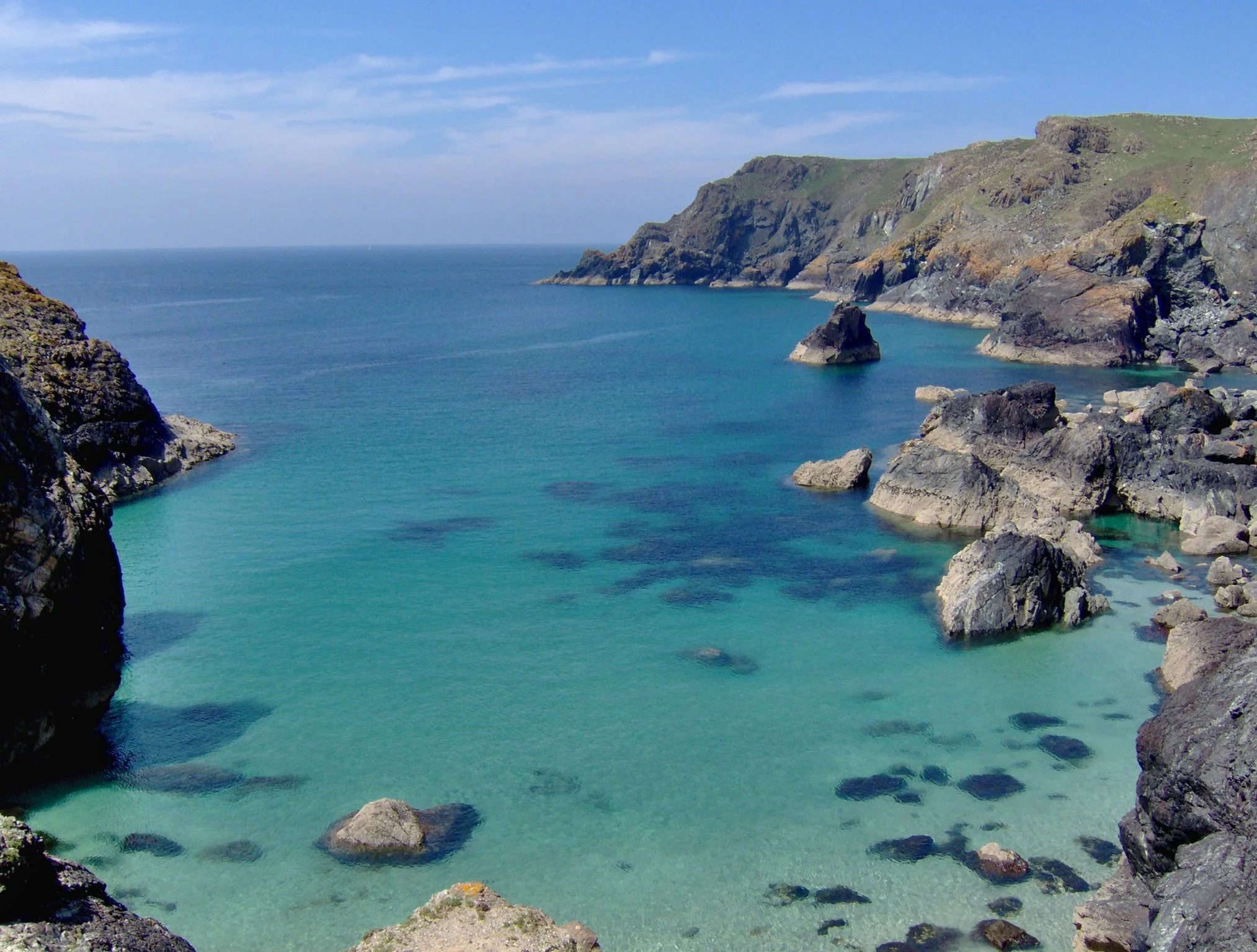 Cliffs overlooking a calm turquoise bay with rocks and clear water, under a partly cloudy sky.