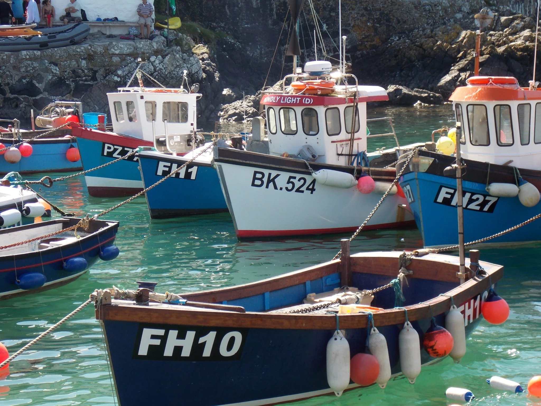 Several boats are docked in Coverack harbour near rocky cliffs. People are sitting and relaxing above, with some equipment and an inflatable boat visible.