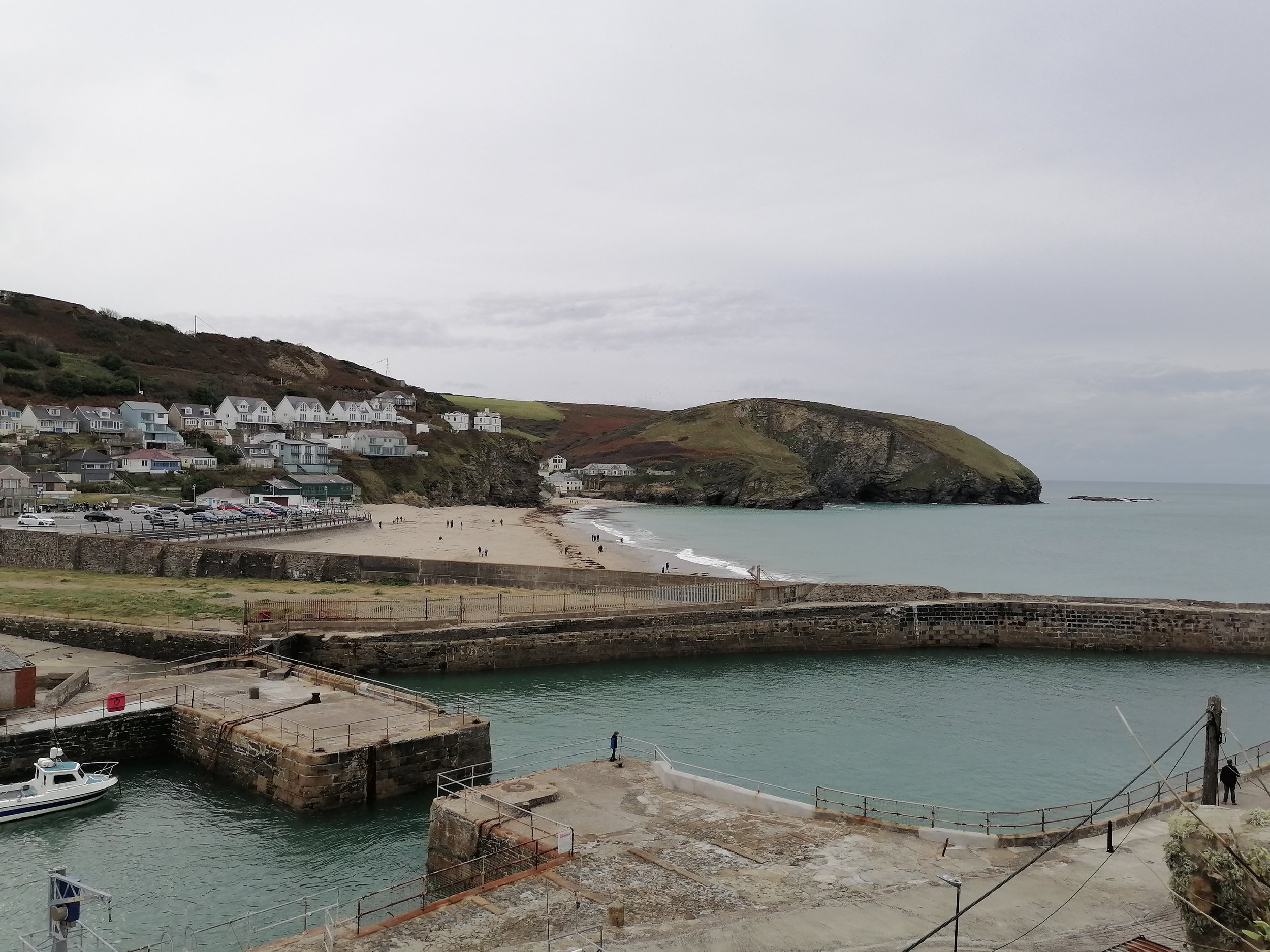 A coastal scene of Portreath Harbour, village and beach with houses on a hillside, a beach with people, and a harbor with boats and a dock. Sunny sky and rocky cliffs in the background.
