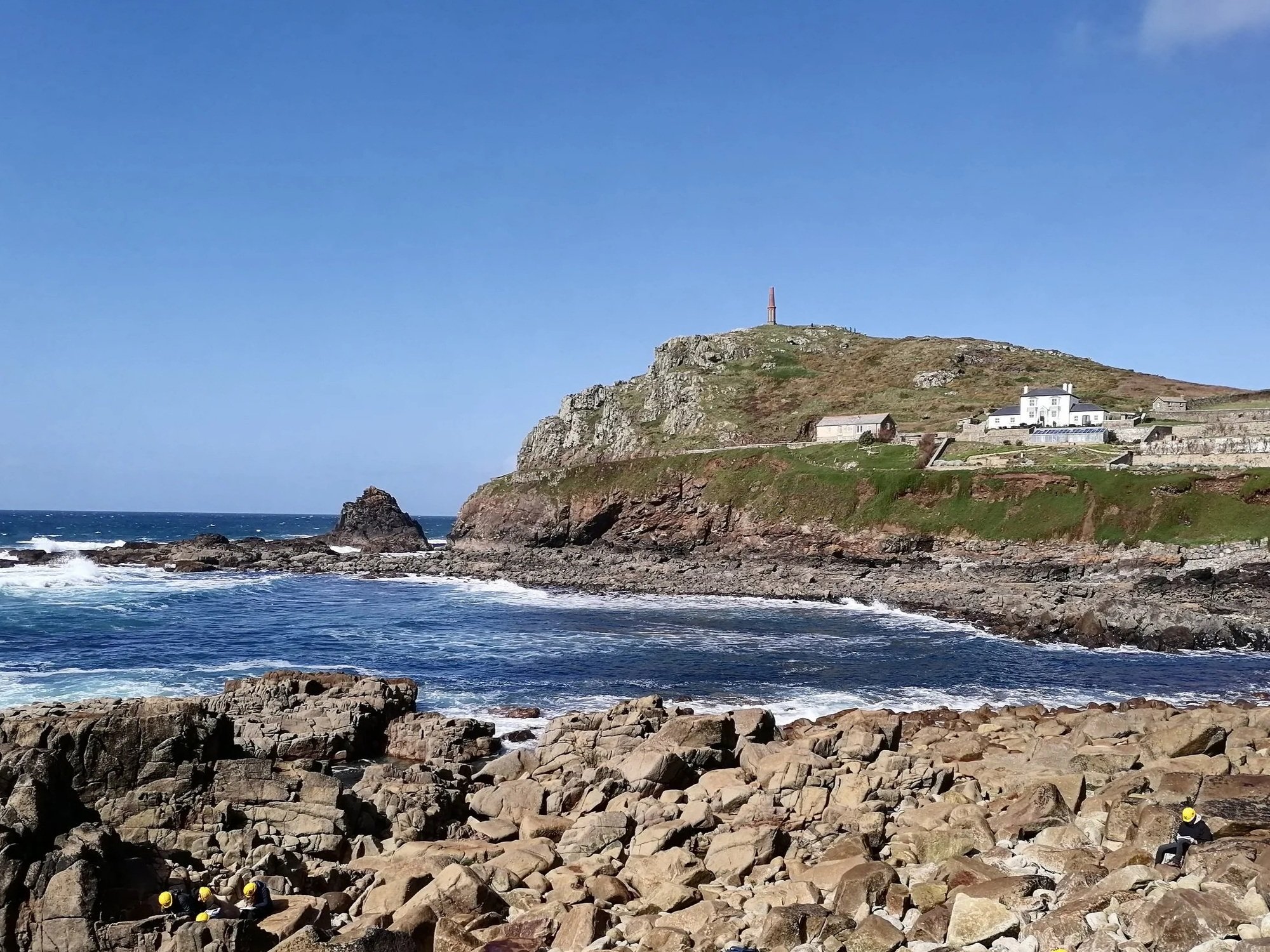 Coastal scene at Cape Cornwall with rocky shoreline, waves crashing, and steep grassy hill with white buildings and monument on top, under a blue sky with some clouds.