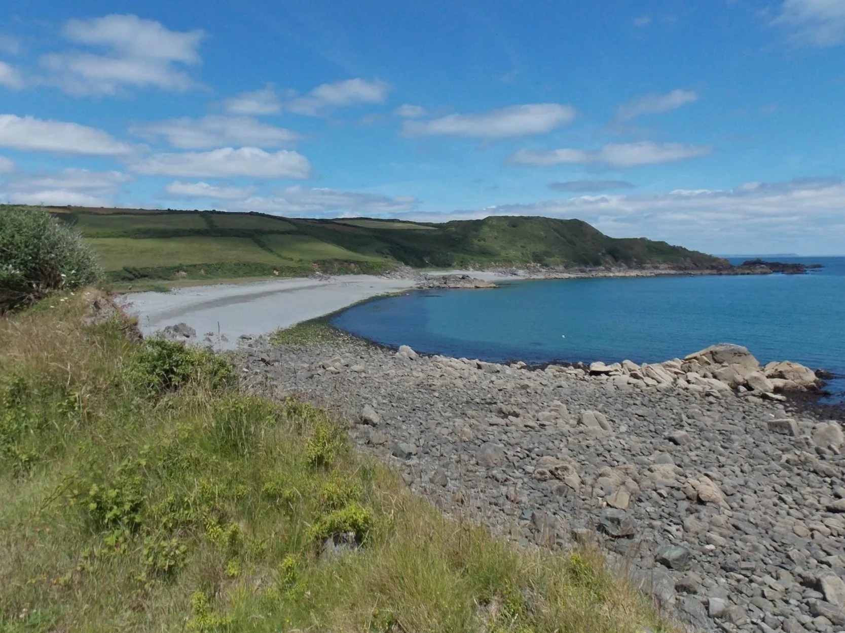 Scenic view Godrevy Cove with green hills in the background, under a blue sky with scattered clouds.