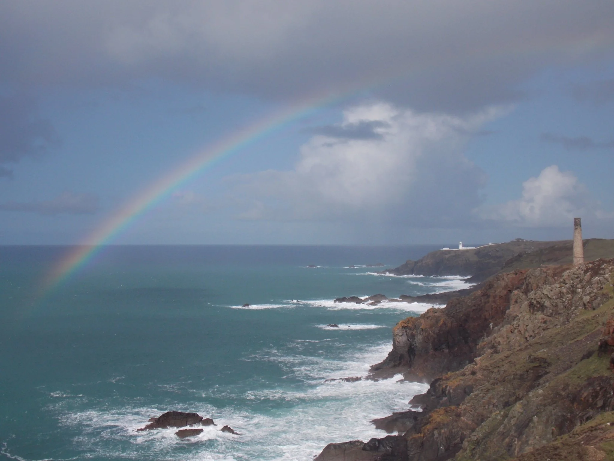 A coastal scene at Levant Mine with rocky cliffs, the ocean with waves crashing, a cloudy sky, a faint rainbow, and a lighthouse on the land.