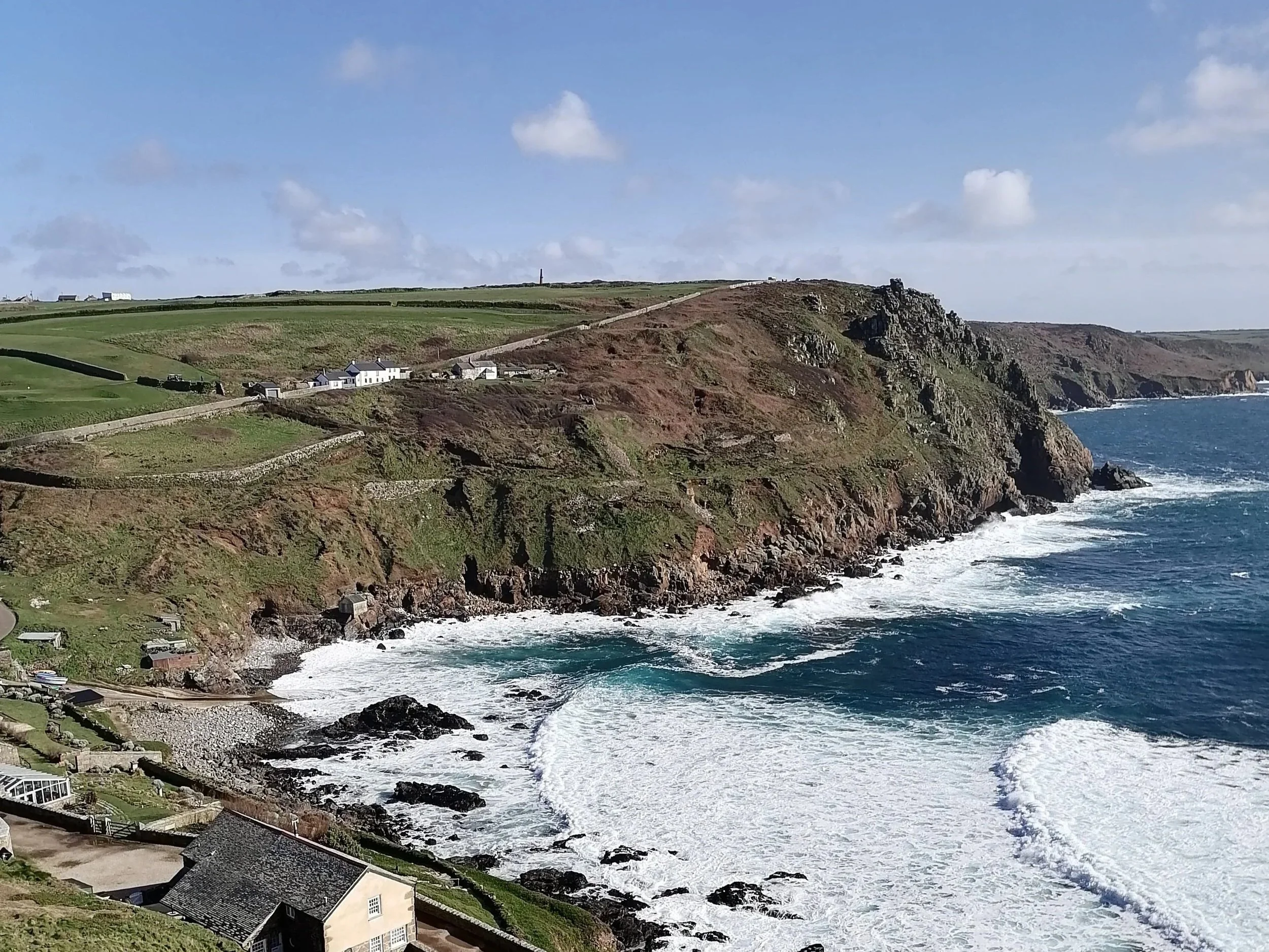 Cliffside view of Priest's Cove from Cape Cornwall with rocky shores, waves crashing below, and a few houses on the land. Green fields and a hill  under a partly cloudy sky.