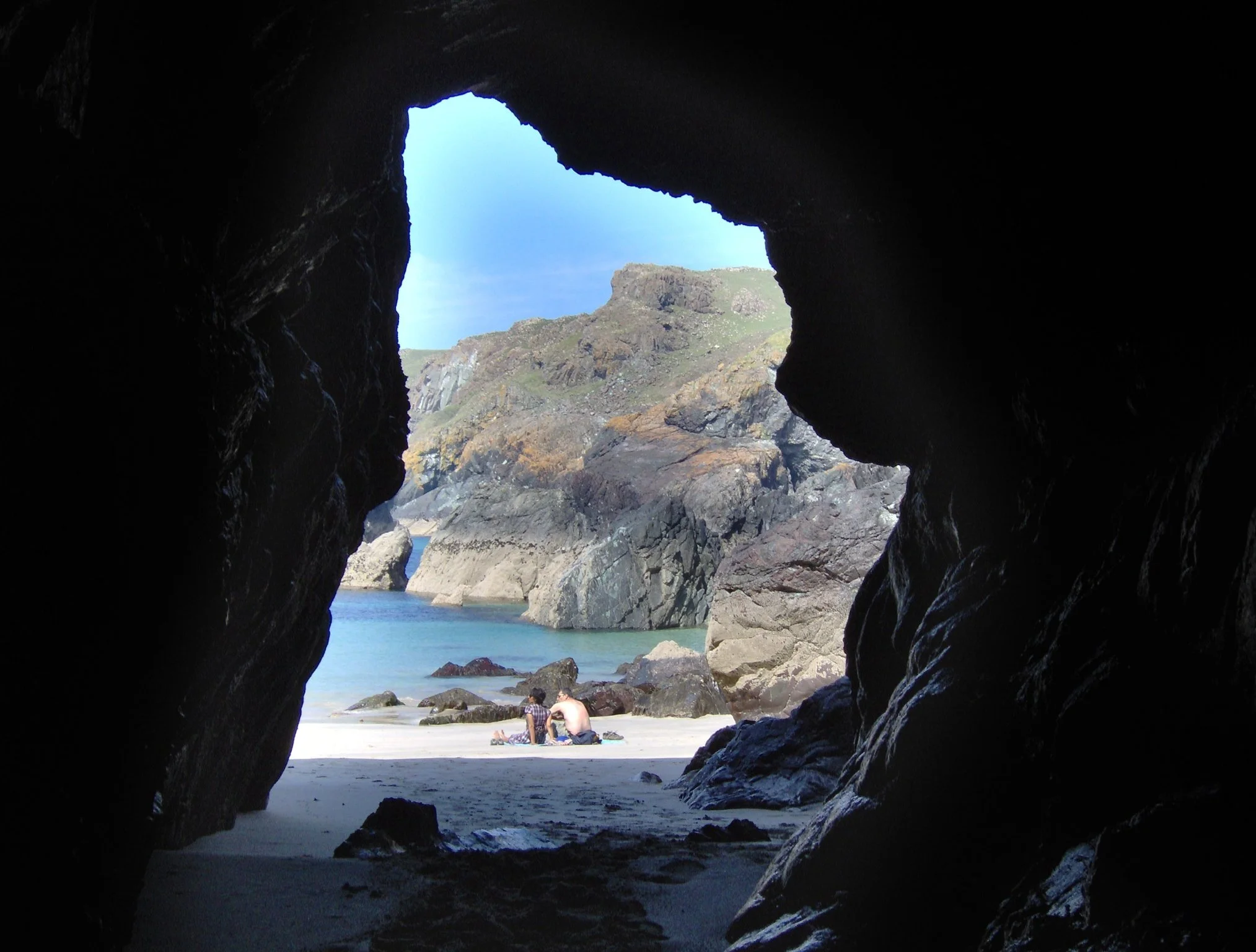 A view of the beach taken from inside a cave, showing rocky cliffs, blue sky, and two people sitting on the sand.