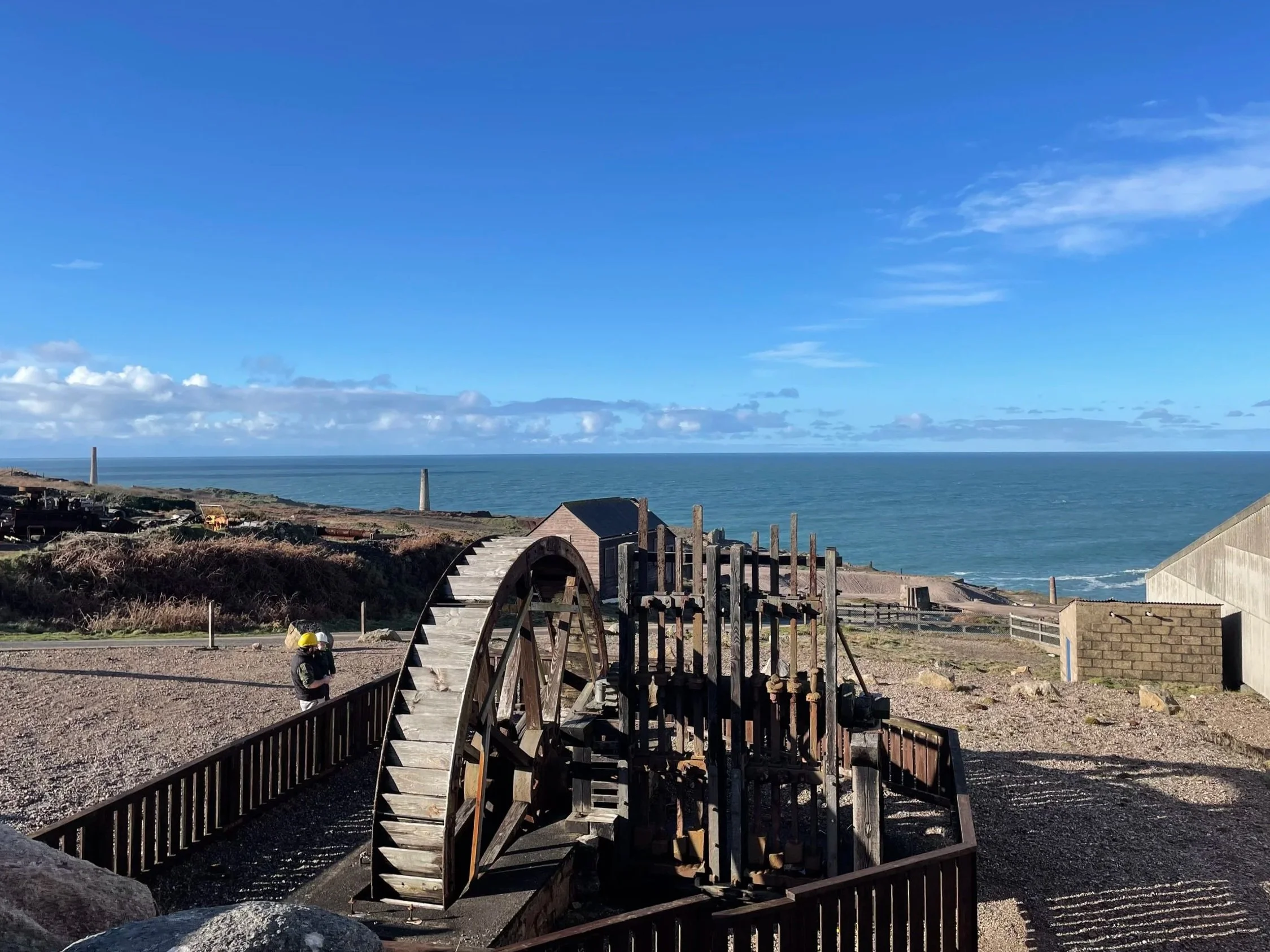 A waterwheel powering a set of Cornish Stamps at Geevor, overlooking the ocean under a partly cloudy sky.